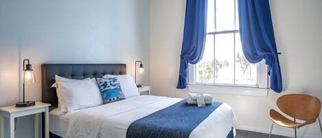 Een net opgemaakt bed met een blauwe deken, een bedlampje en een stoel in een kamer met een raam en gordijnen.