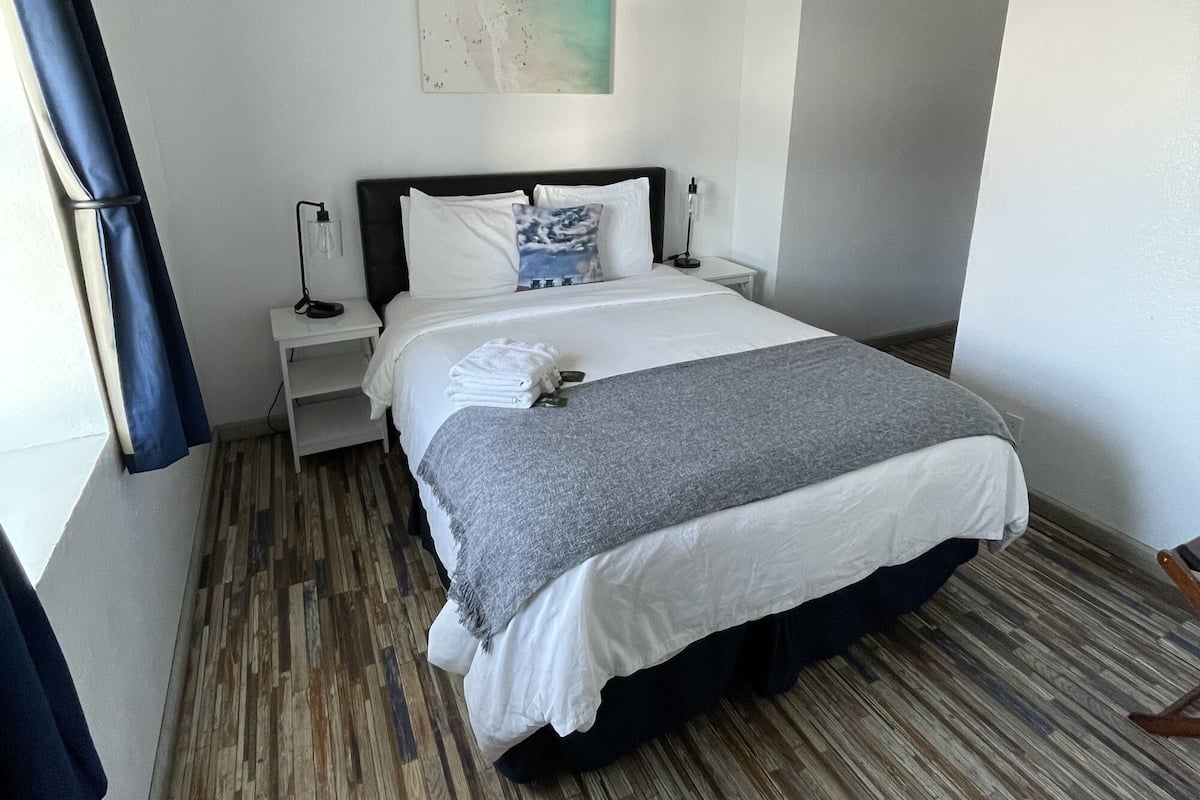Une chambre d’hôtel comprenant un lit, un ventilateur de plafond, une table de chevet, une œuvre d’art accrochée au mur et une fenêtre avec des rideaux.