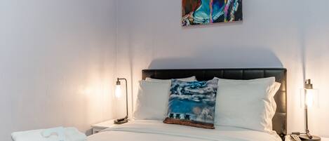 A neatly made bed with white linens, a pillow with a cloud design, and a bedside table with a lamp.
