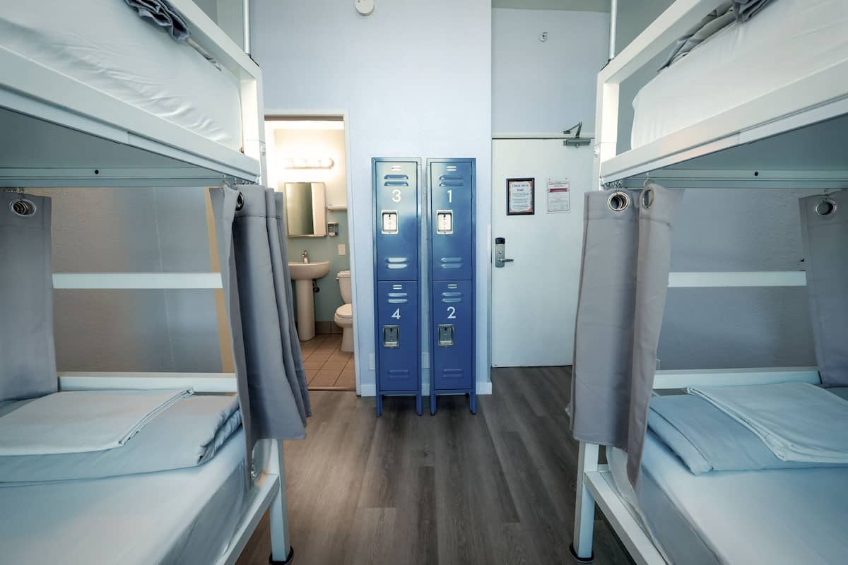 Une chambre d’hôtel avec des lits superposés, une salle de bain avec un lavabo et des toilettes, et des casiers de rangement bleus.