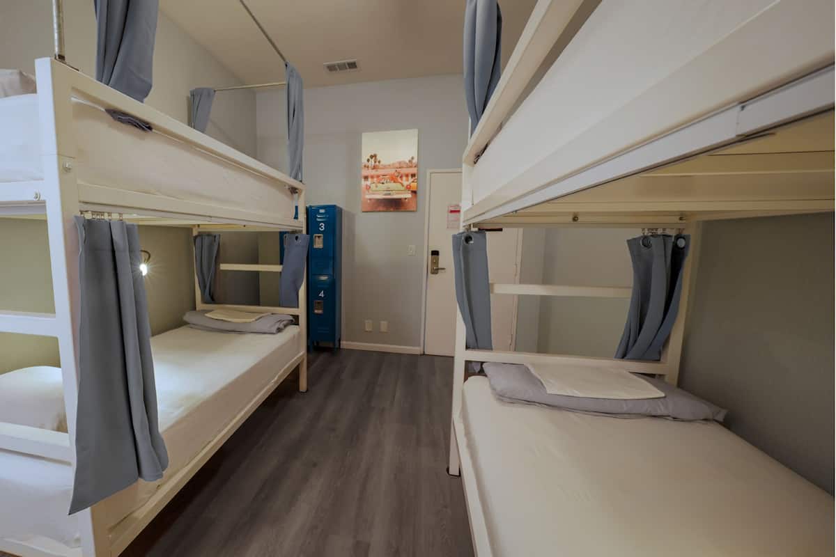 Une chambre de dortoir avec des lits superposés, un meuble de rangement bleu et une fenêtre donnant sur un immeuble.