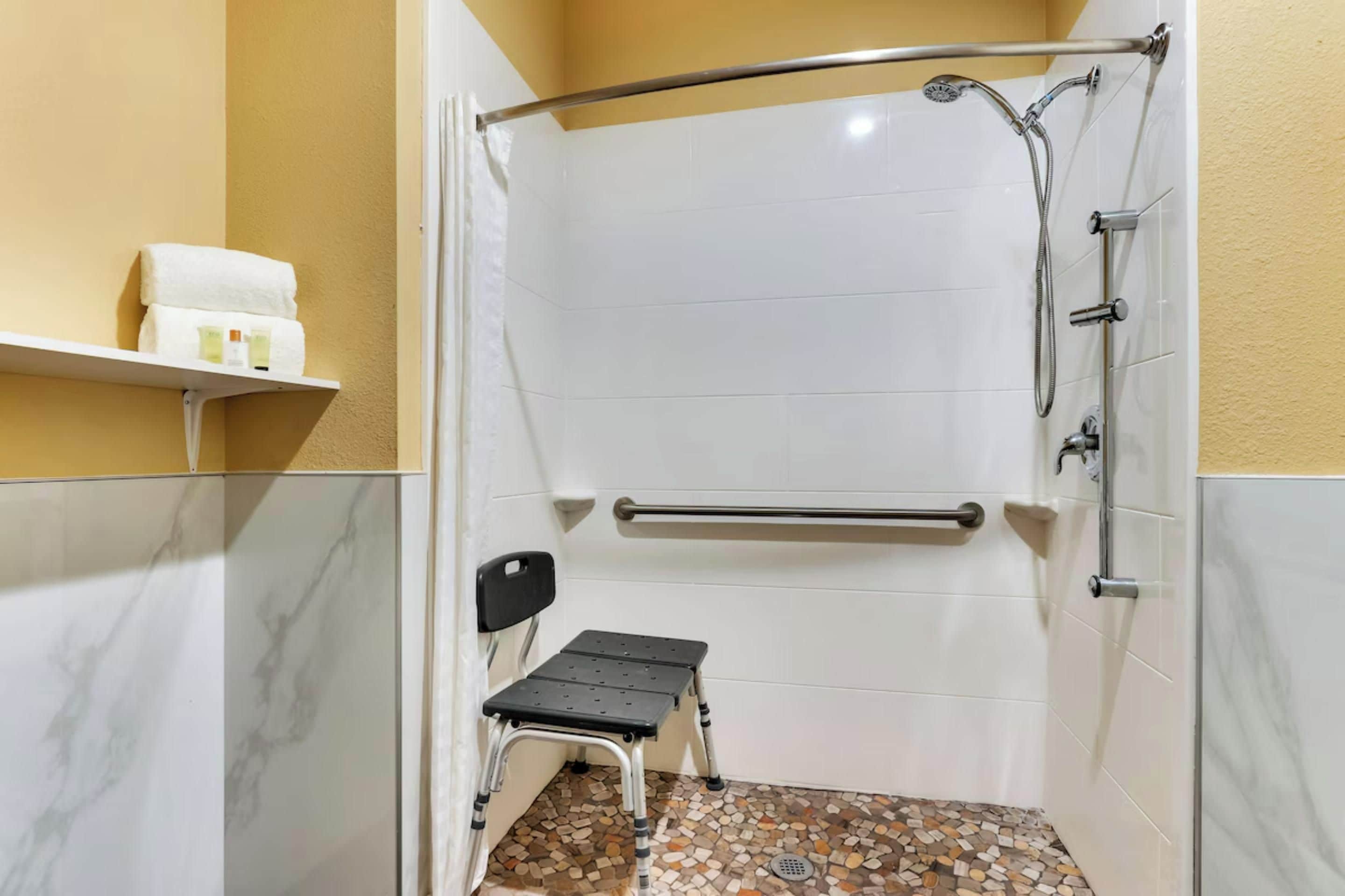 A small, enclosed shower area with a grab bar, a step stool, and a shelf with toiletries.
