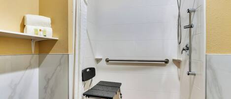 A small, enclosed shower area with a grab bar, a step stool, and a shelf with toiletries.