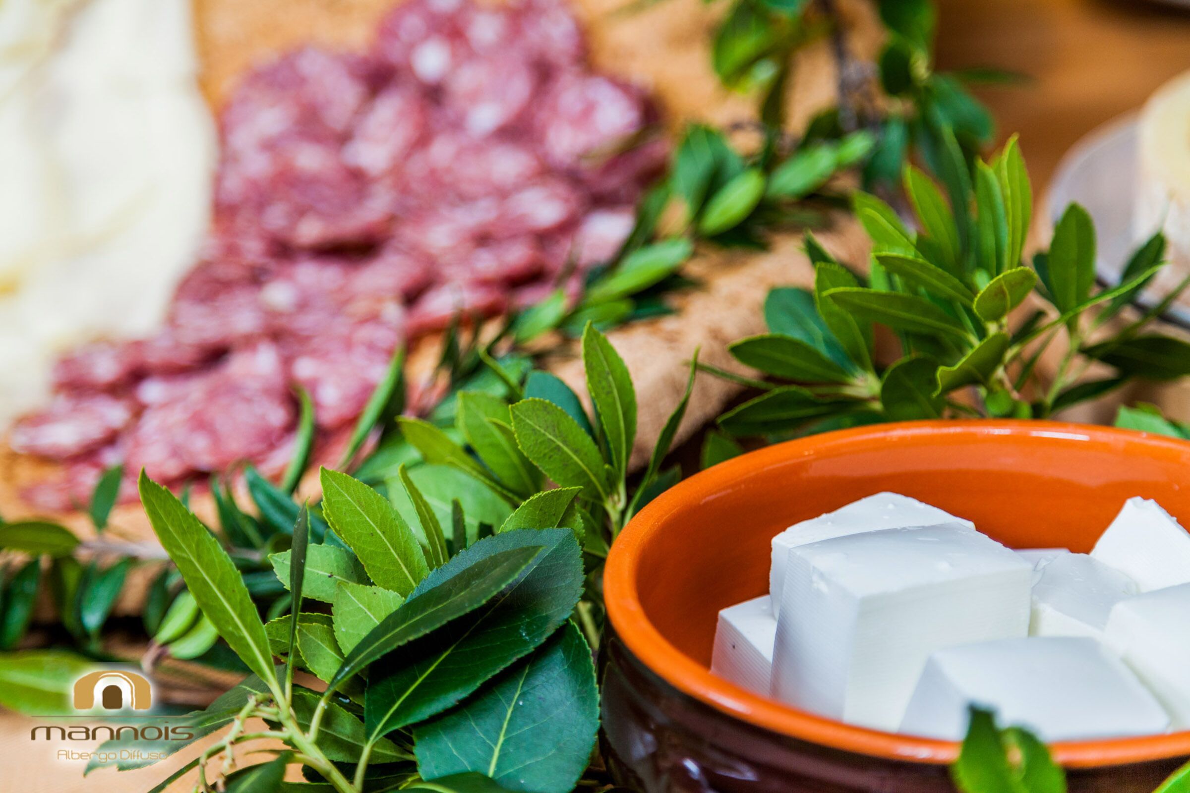 A bowl of white cubes, possibly cheese, surrounded by green leaves and a plate of sliced meats.