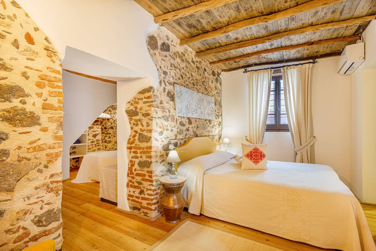A bedroom with a stone wall, wooden beams, a bed with white bedding, a bedside lamp, and a window with curtains.