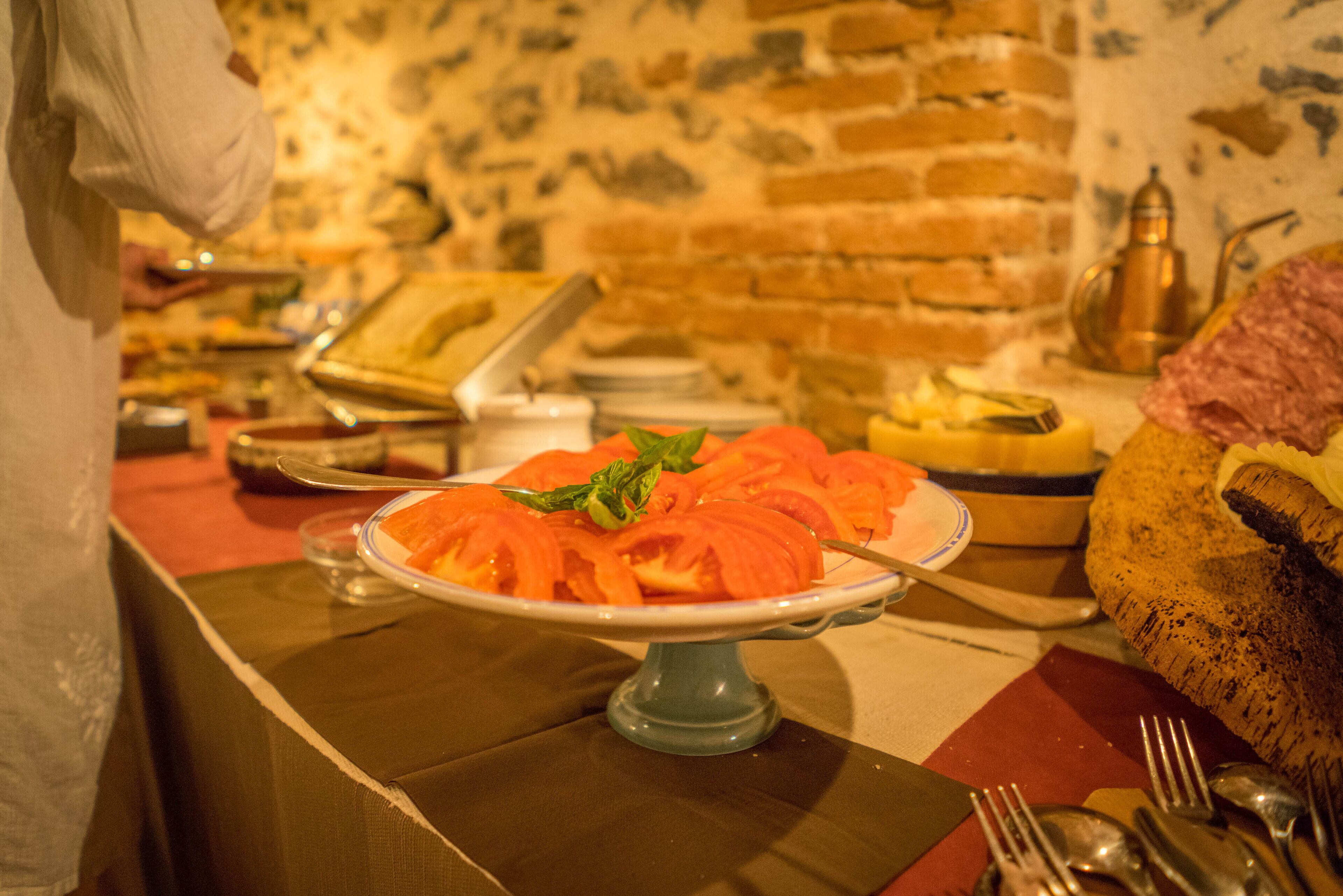 A buffet with various dishes, including a plate of sliced tomatoes garnished with basil, set against a brick wall background.