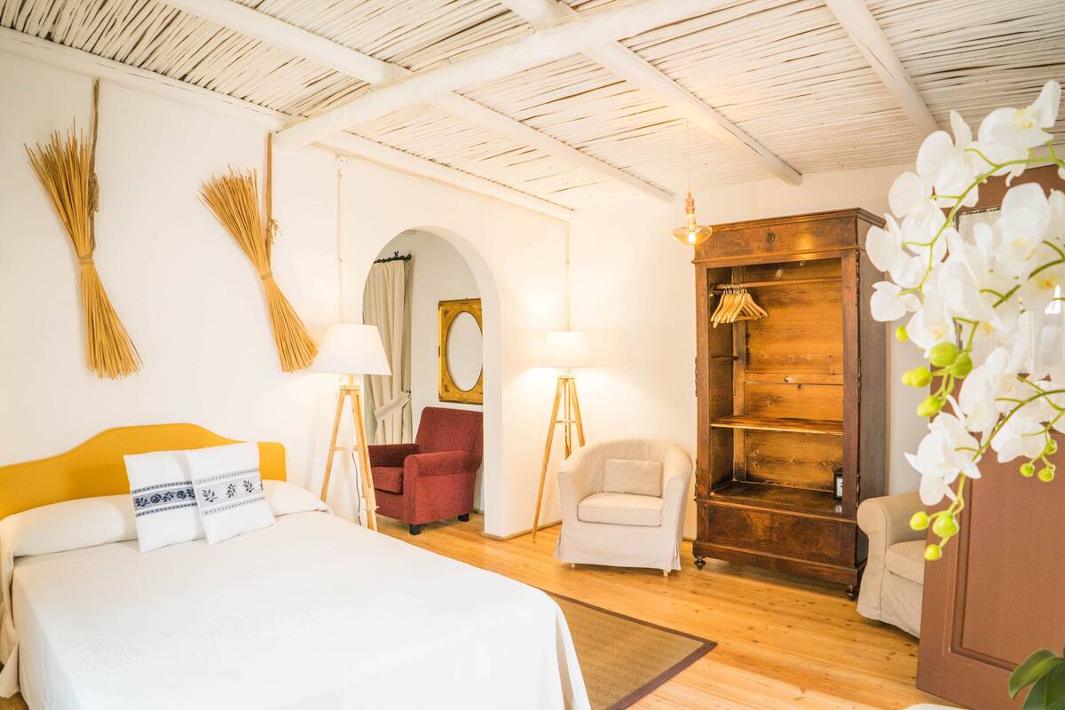 A hotel room with a bed, a red armchair, a white armchair, a wooden wardrobe, and a mirror.