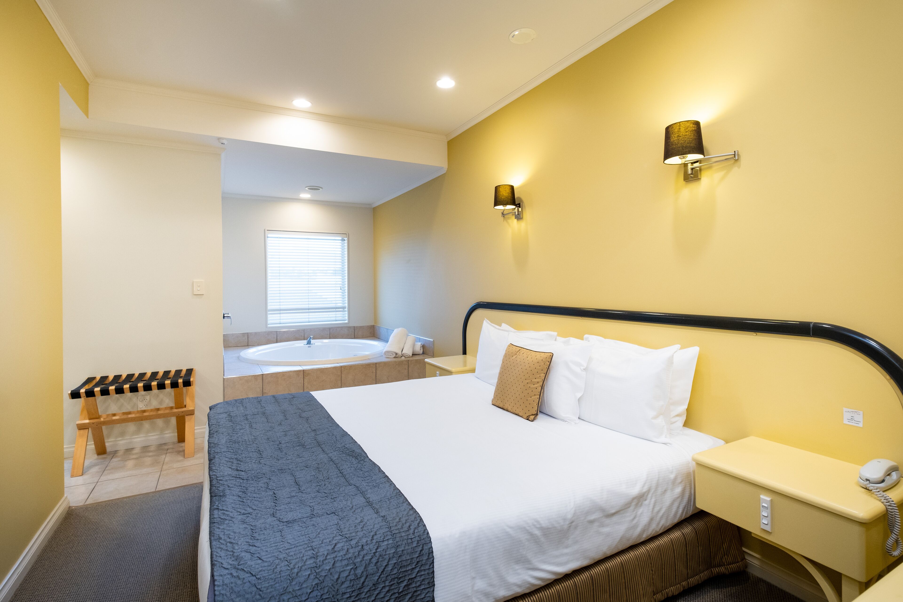 A hotel room with a large bed, a bedside table with a telephone, a bench, a bathtub, and a window with blinds.