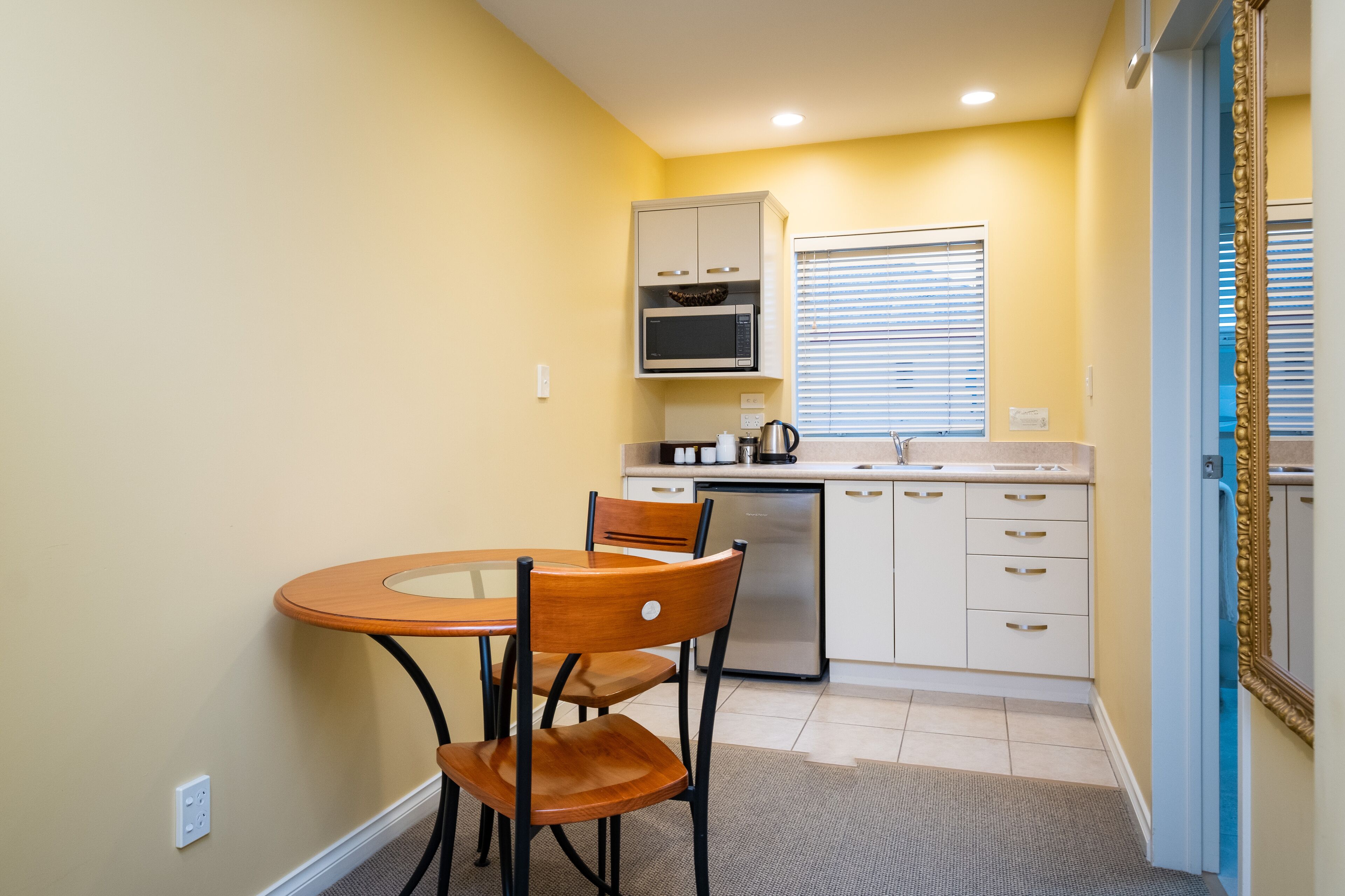 A compact kitchen with a microwave, stove, and sink. There is a dining table with chairs and a window with blinds.