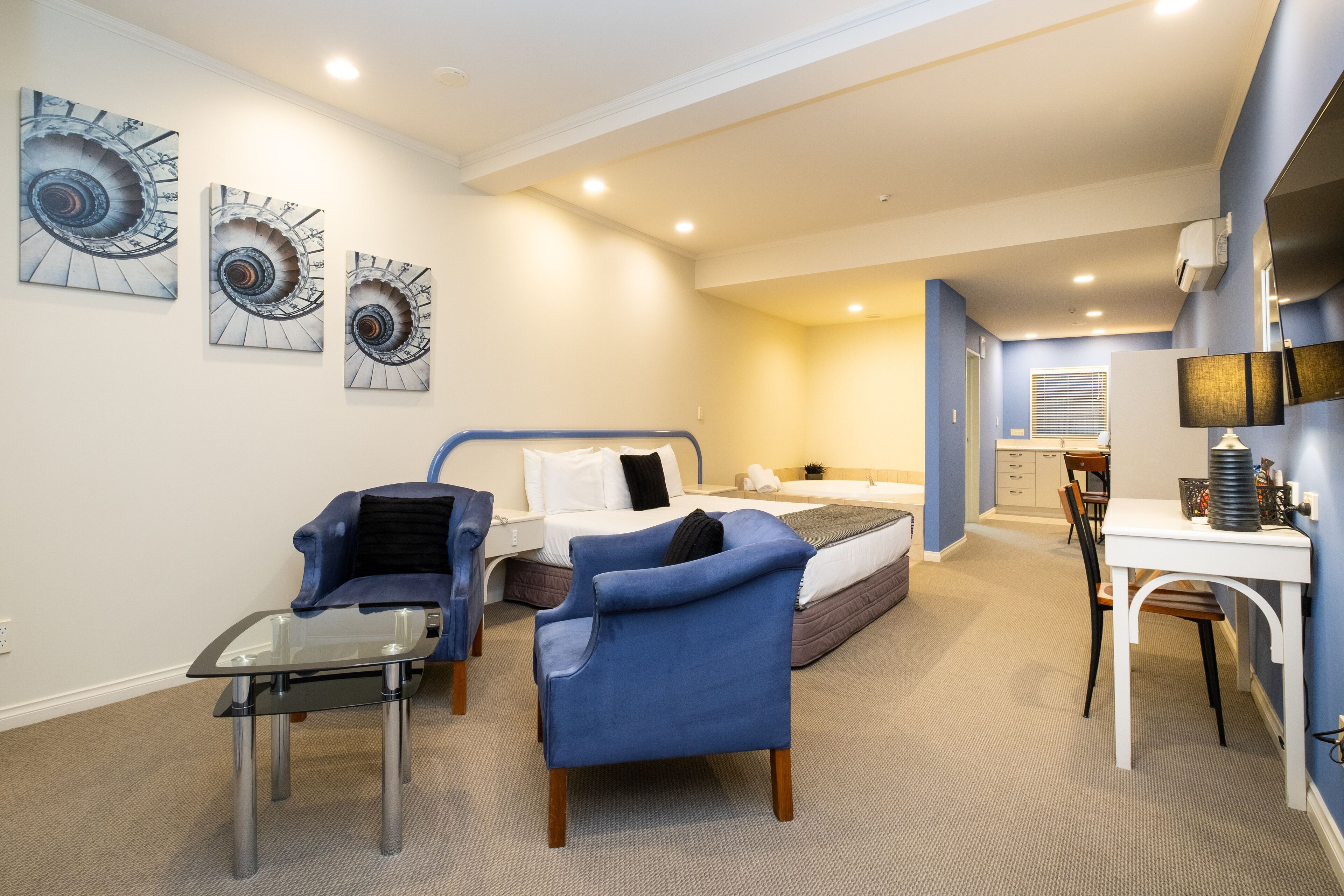 Habitación de hotel con una cama, dos sillones azules, una mesa de centro de vidrio, un escritorio con una lámpara y tres obras de arte espirales en la pared.