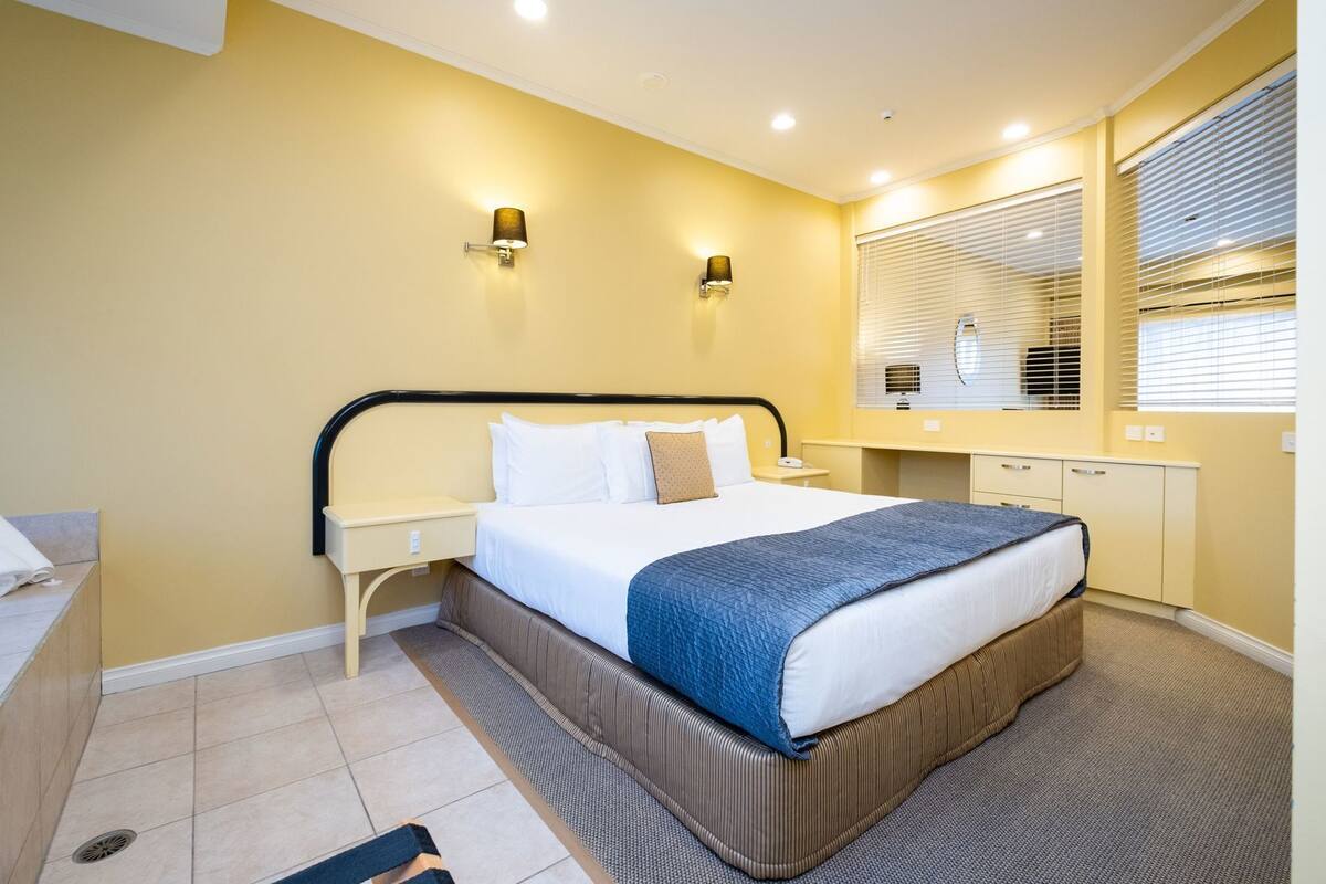 A hotel room with a bed, bedside tables, a desk, and a window with blinds.