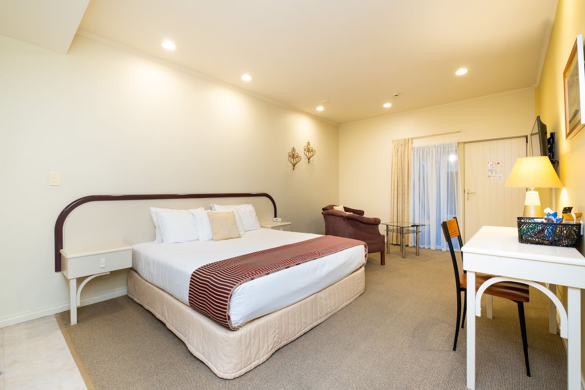 A hotel room with a bed, bedside table, chair, desk, lamp, and curtains.