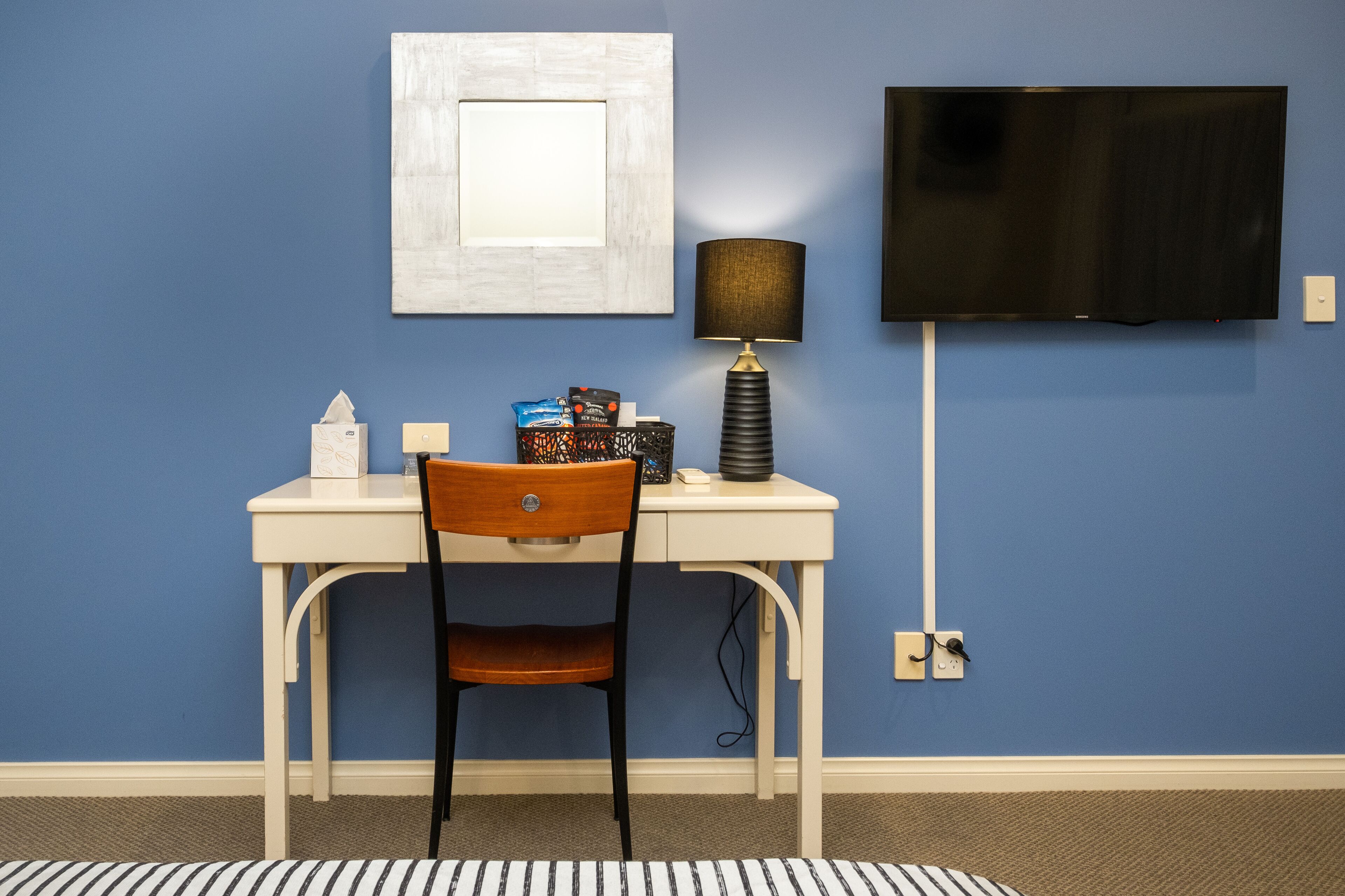 Una habitación con pared azul, un escritorio blanco, una silla de madera, un televisor montado en la pared y una lámpara sobre el escritorio.
