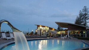 Una piscina con cascada, sillas de descanso y un edificio moderno con grandes ventanales.