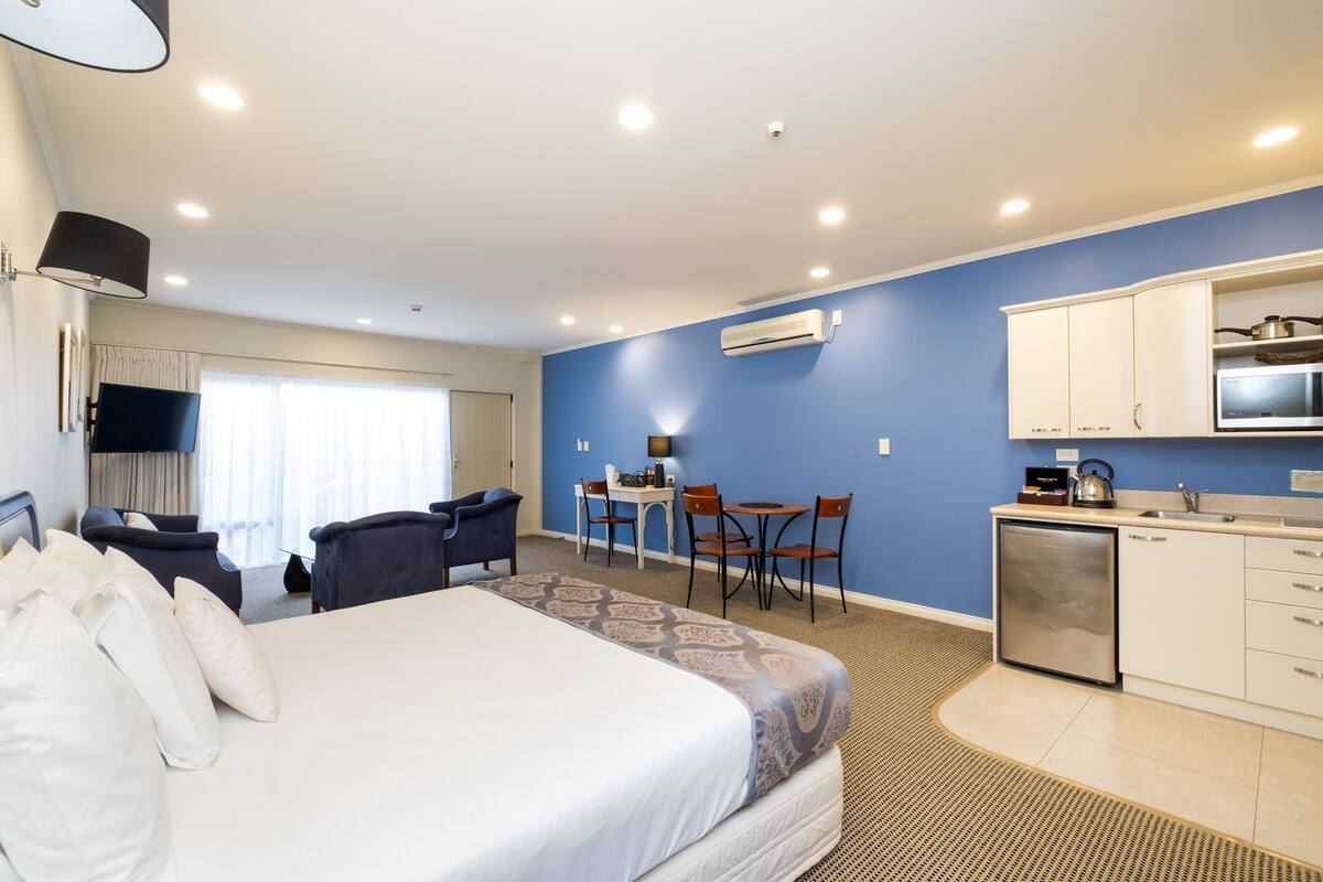 A hotel room with a bed, a desk, chairs, a kitchenette, and an air conditioning unit.