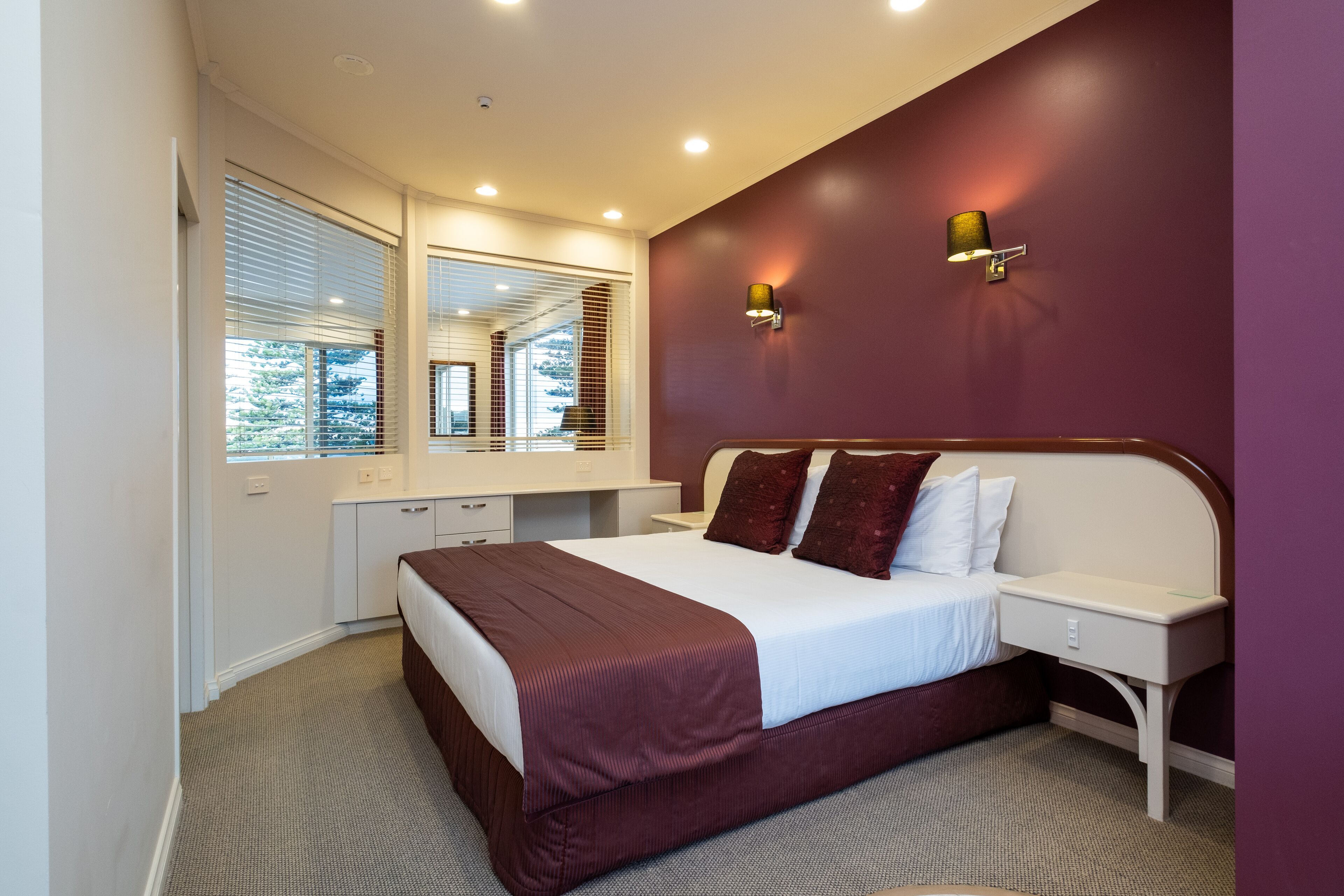 A modern bedroom with a large bed, a bedside table, a mirror, and a window with blinds.