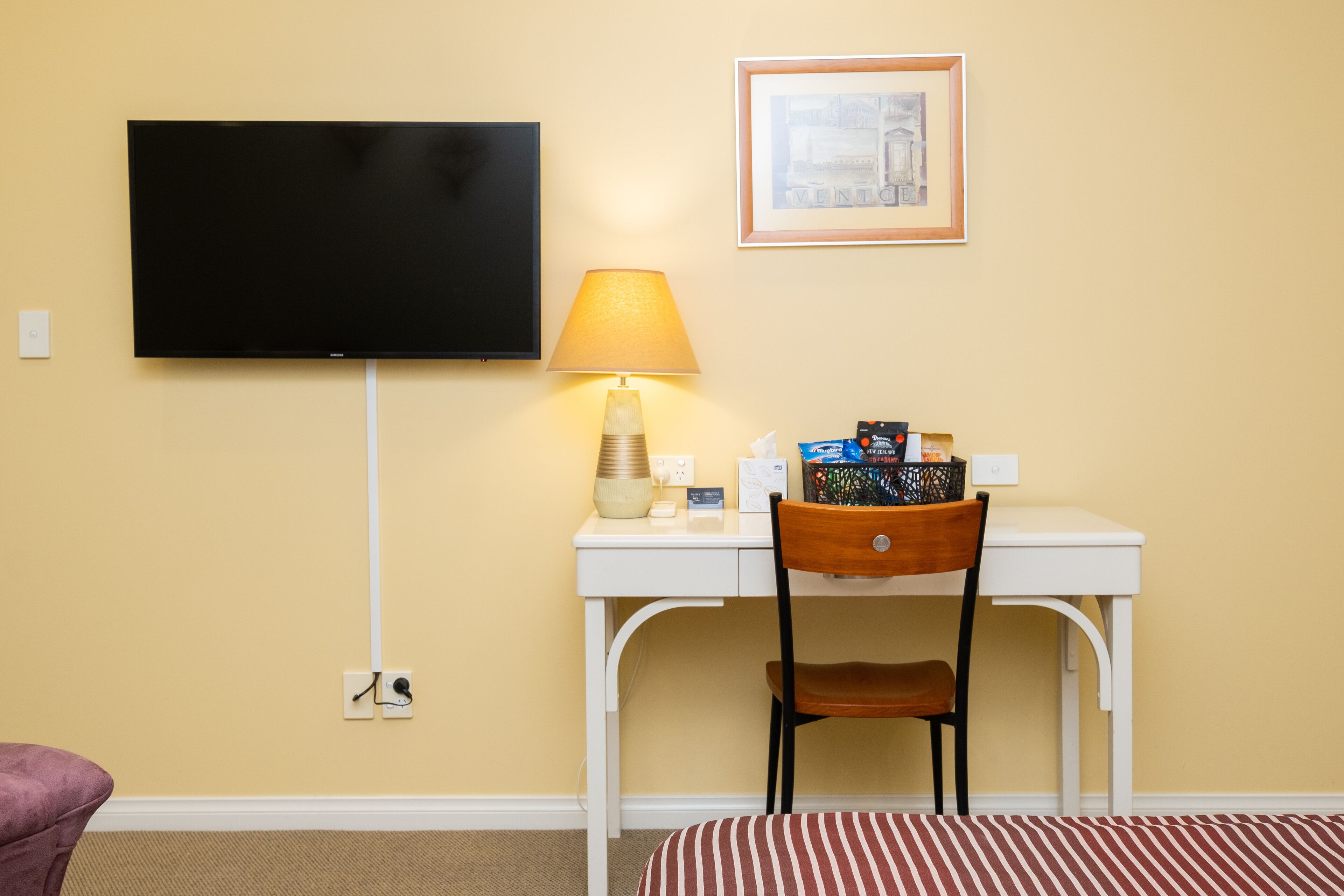 Een kamer met een aan de muur bevestigde televisie, een bureau met een lamp, een stoel en een lijst met een afbeelding.