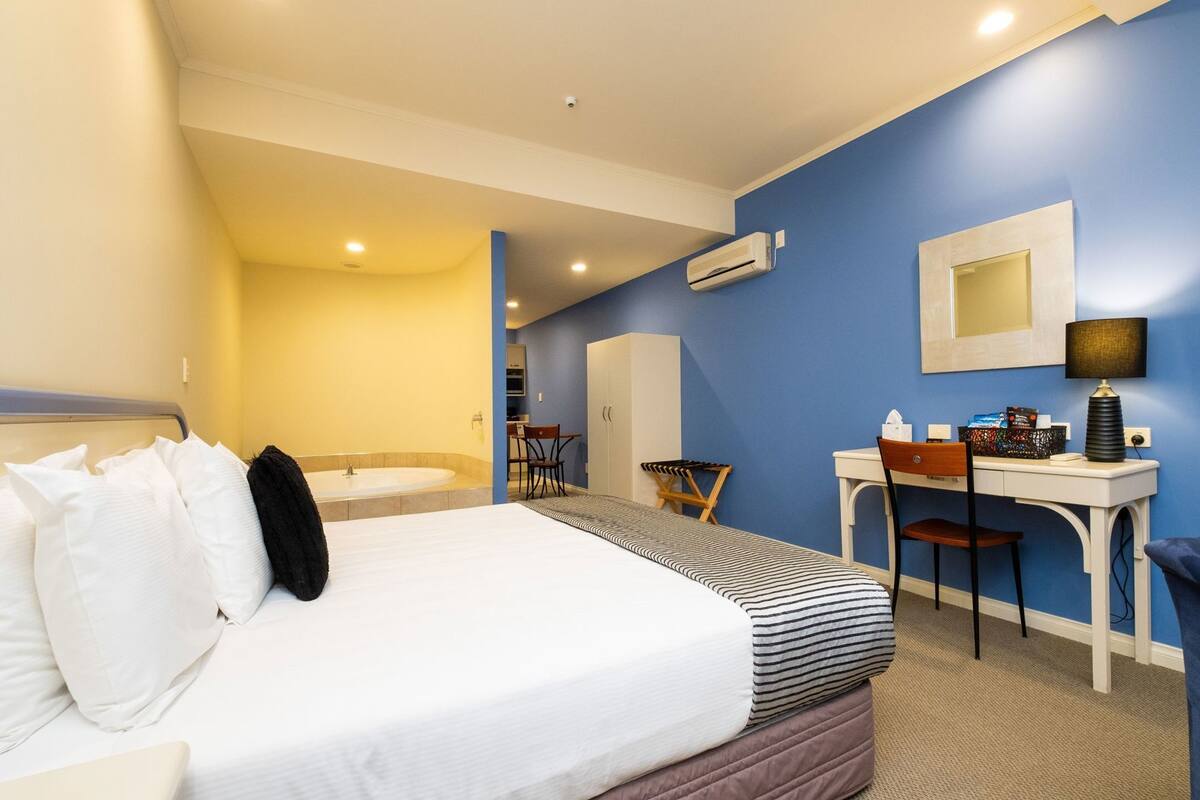 A hotel room with a bed, a desk, a chair, a small table, a mirror, and an air conditioning unit.