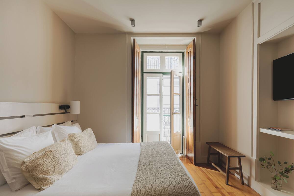 Un dormitorio con cama, lámpara de noche, banco de madera y televisor sobre una estantería.