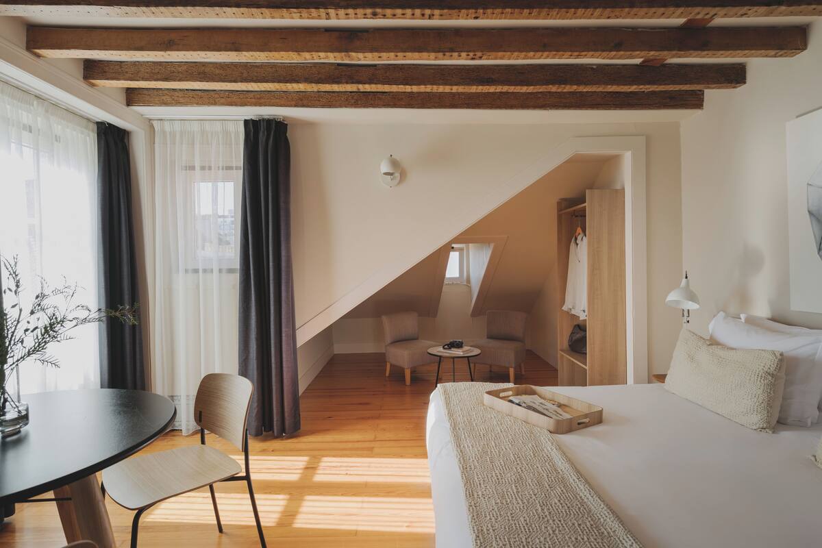 Una habitación acogedora con una cama, un escritorio y una silla. Hay una mesita con una lámpara y una planta. La habitación tiene vigas de madera en el techo y una ventana con cortinas.