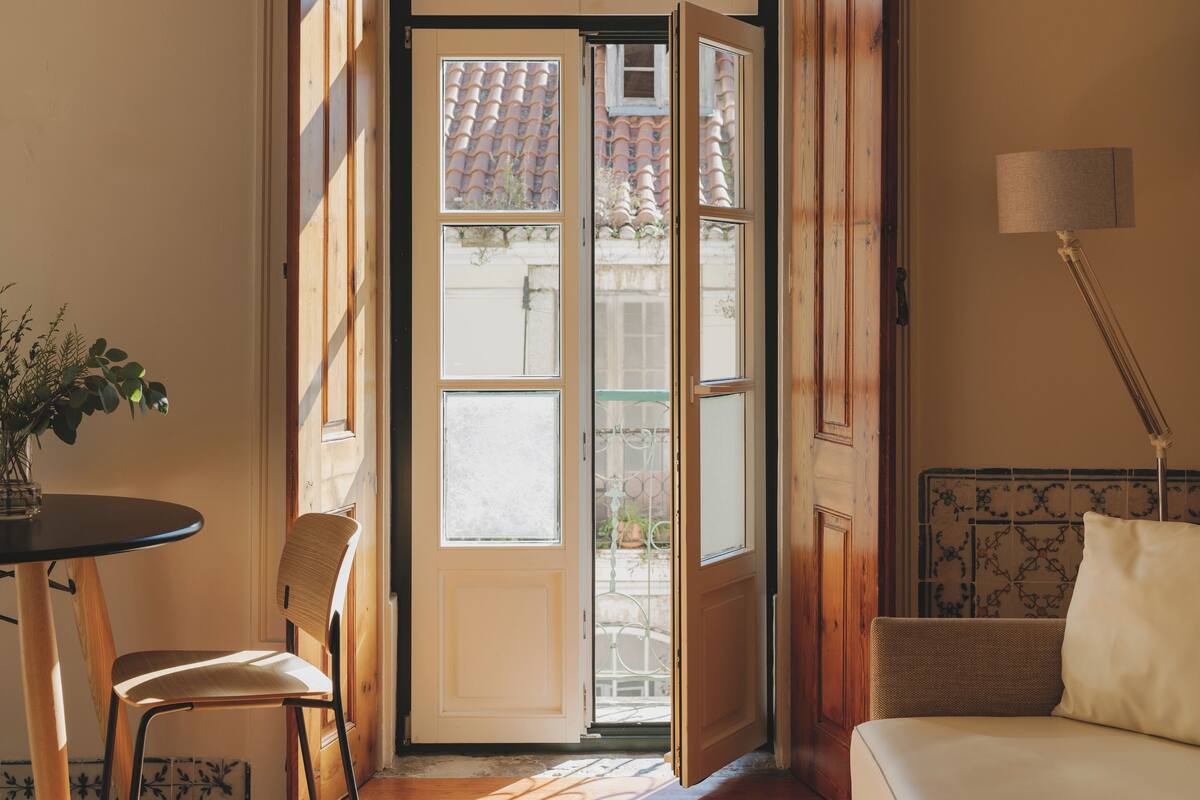 Una habitación con piso de madera, un sofá blanco, una mesa de comedor con una silla y un ventanal con vista a un tejado de tejas.