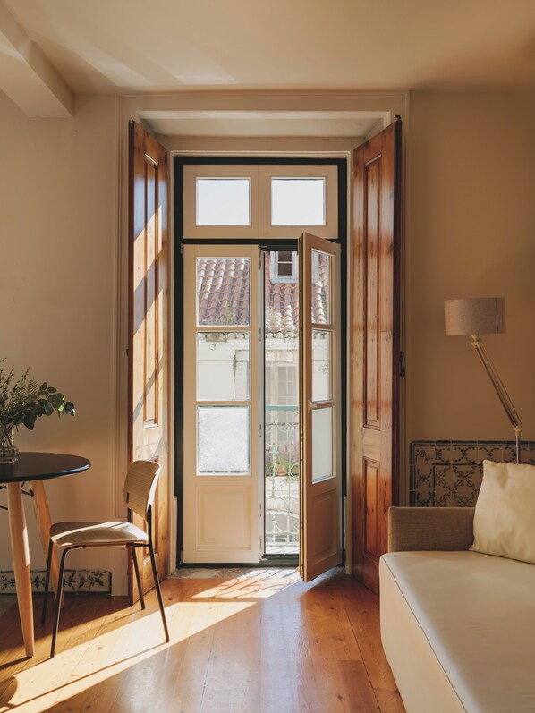 Une pièce avec un plancher en bois, un canapé blanc, une table à manger avec une chaise, et une grande fenêtre donnant sur un toit recouvert de tuiles.