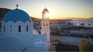 A coastal town with a prominent white church featuring a blue dome and a bell tower, set against a backdrop of a sunset over the sea. - Aquarius Village Agistri (Agistri)