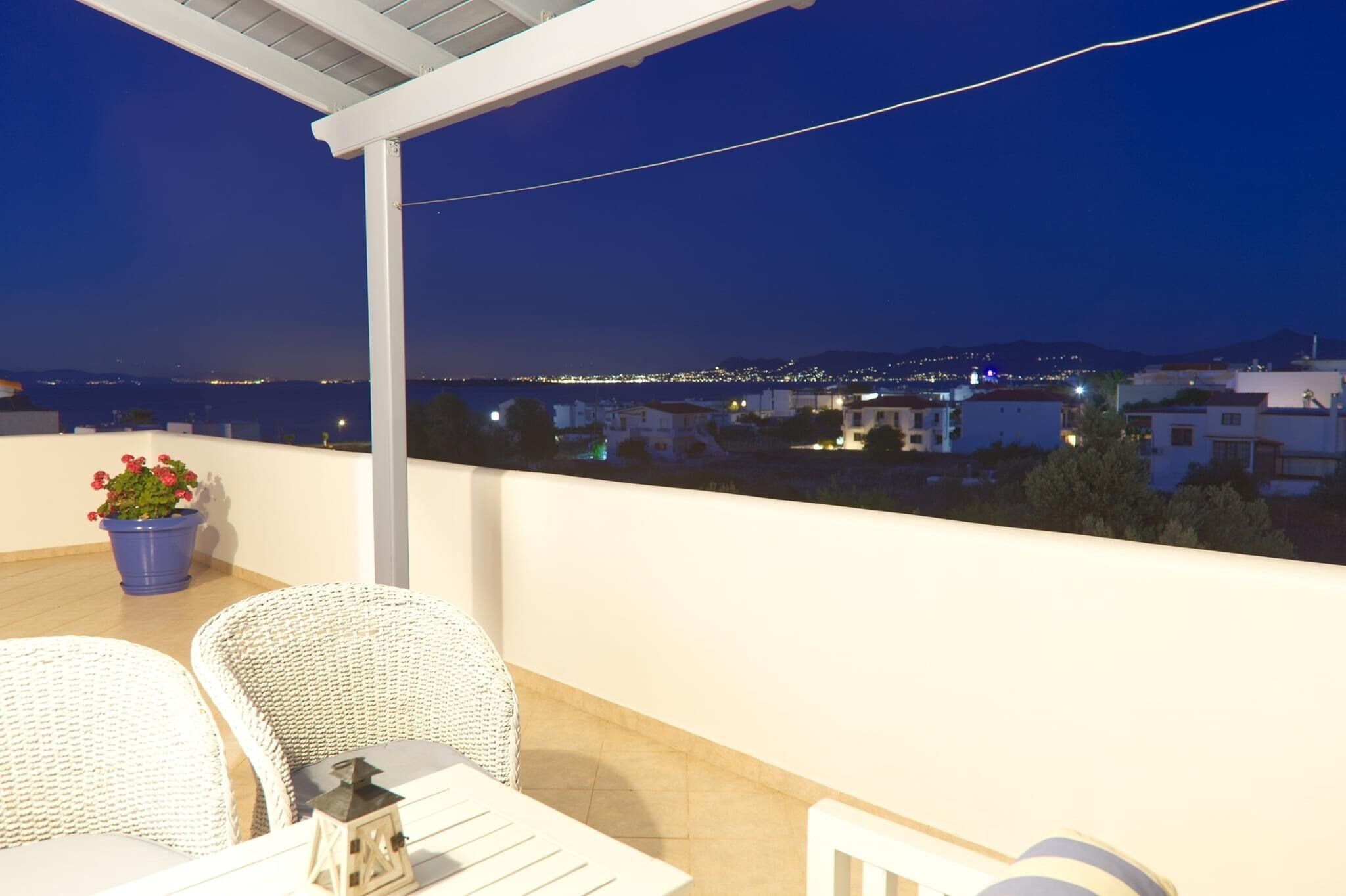 A balcony with wicker chairs and a table, overlooking a cityscape at night.