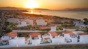A coastal residential area with houses featuring red-tiled roofs and white walls, arranged in a row along a road with a view of the sea and mountains in the background. - Aquarius Village Agistri (Agistri)