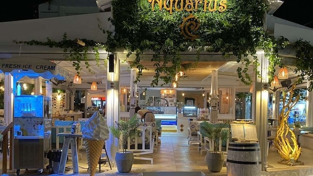 An outdoor entrance to a restaurant with a sign reading "Aquarius." There are tables and chairs outside, and potted plants are placed around the area. - Aquarius Village Agistri (Agistri)