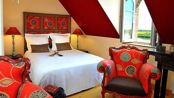 A bedroom with a large bed, two red upholstered chairs, a wooden desk, and a window with a view of greenery.