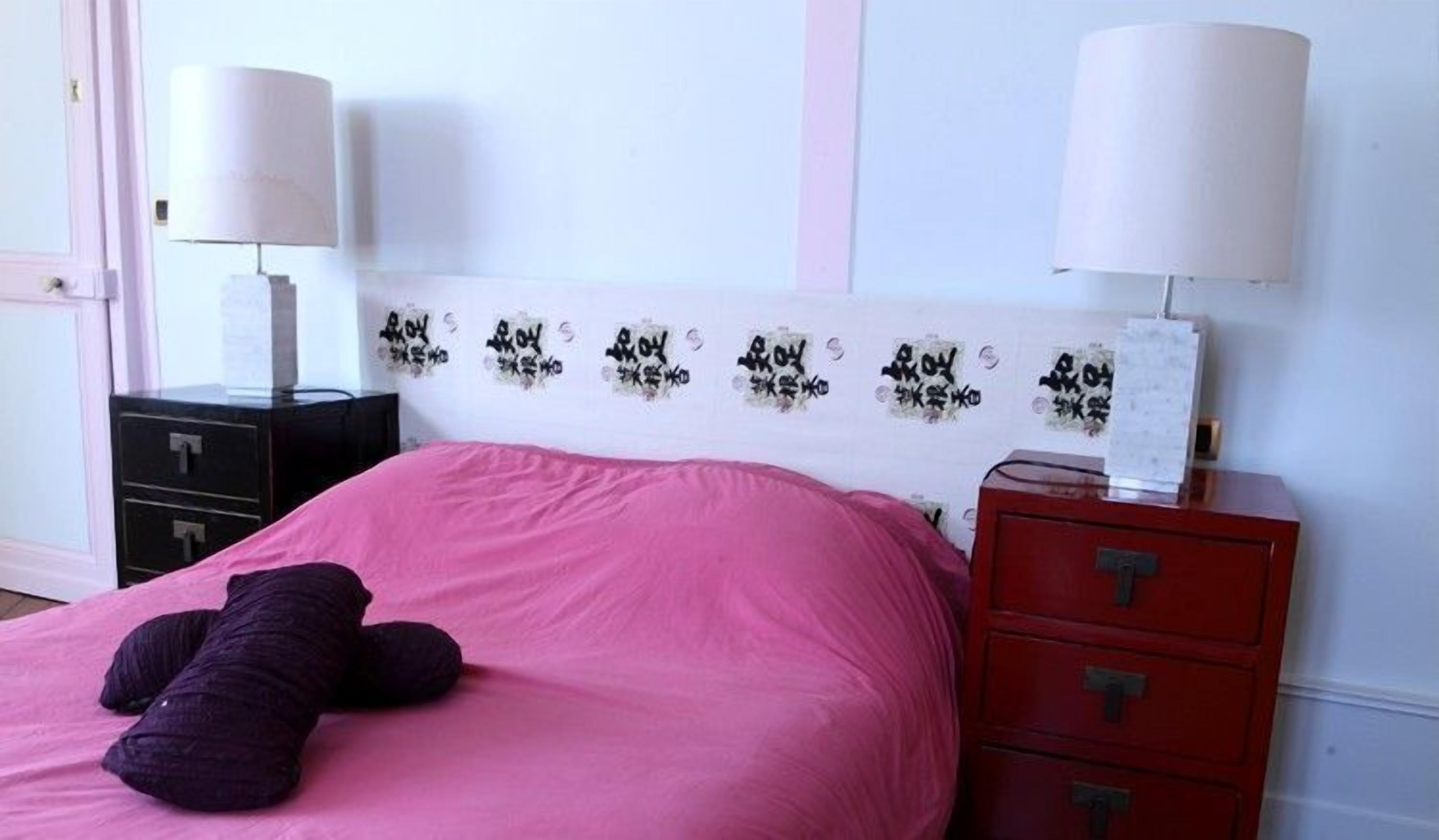 Un dormitorio con una colcha rosa, un peluche morado, un cabecero blanco con texto negro, dos mesitas de noche con lámparas y un aparador rojo.