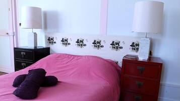 Un dormitorio con una colcha rosa, un peluche morado, un cabecero blanco con texto negro, dos mesitas de noche con lámparas y un aparador rojo.