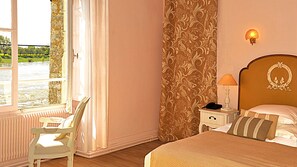A bedroom with a bed, a nightstand, a lamp, a chair, and a view of a river through a window. - Le Relais Louis XI (Meung-sur-Loire)