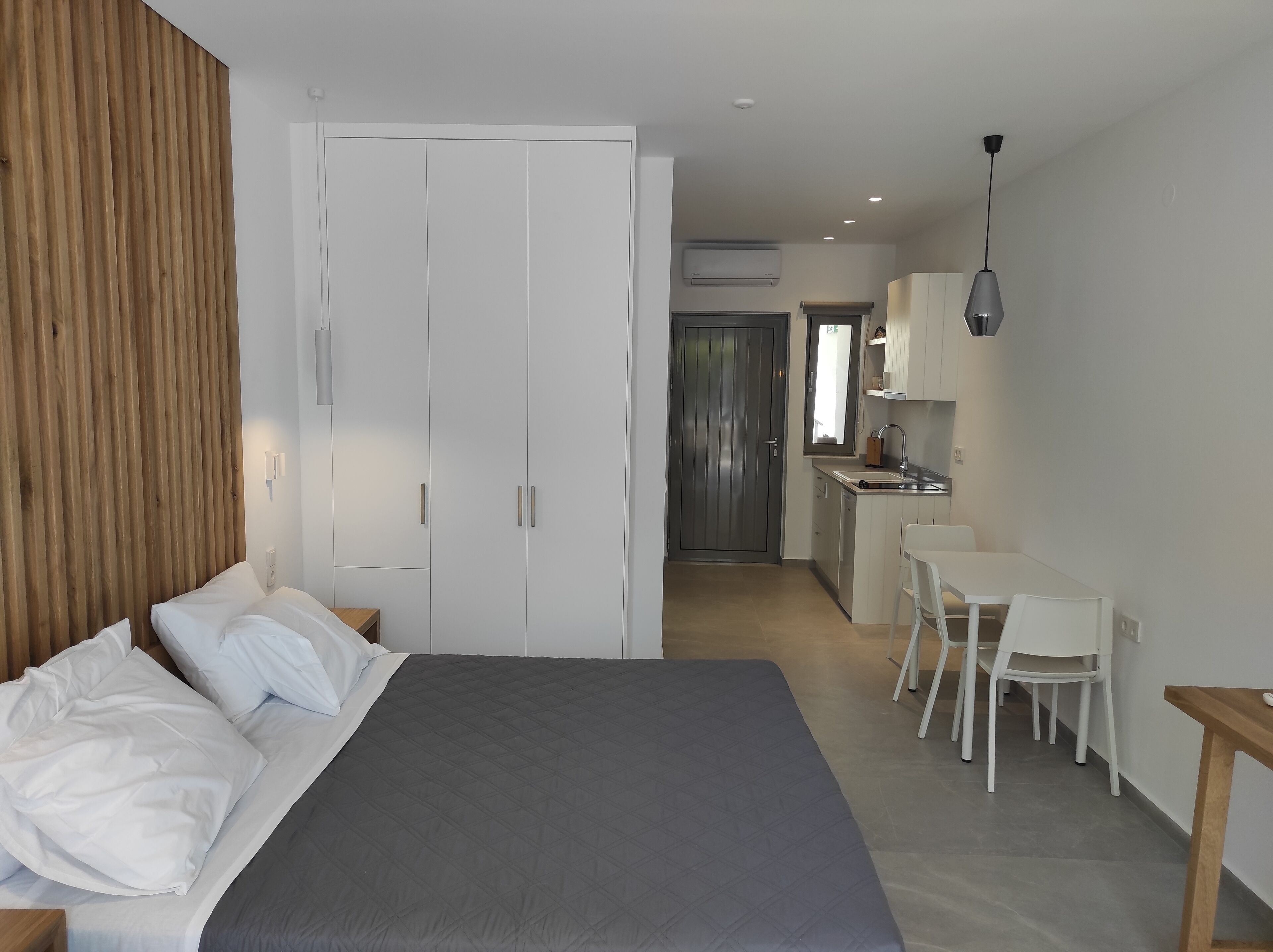 Une chambre moderne avec un grand lit, un mur recouvert de panneaux en bois, une armoire blanche et un coin cuisine comprenant une table et des chaises.