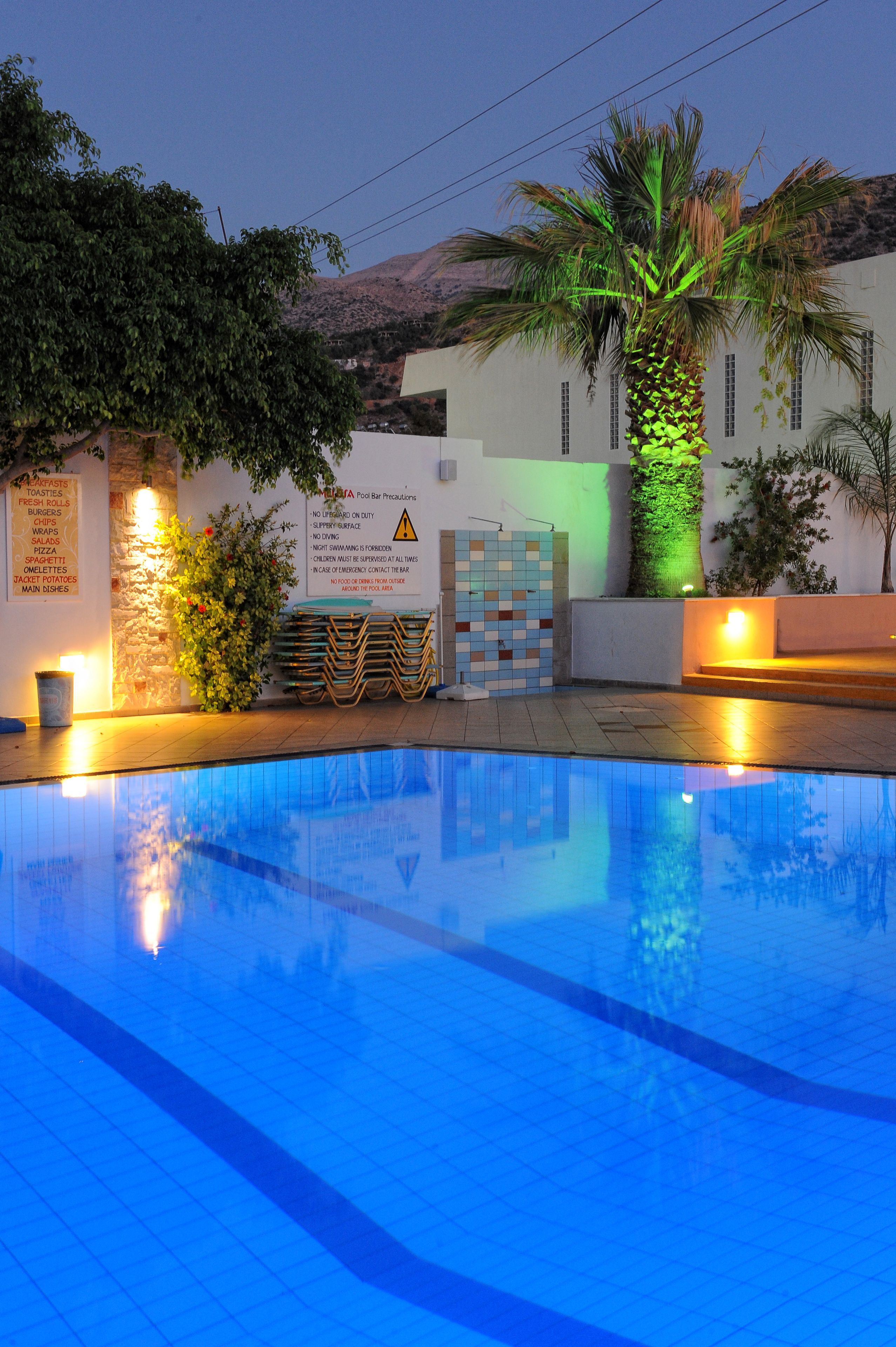 Piscina com deck azulejado, cercada por um edifício com placa indicando opções de comida e bebida.
