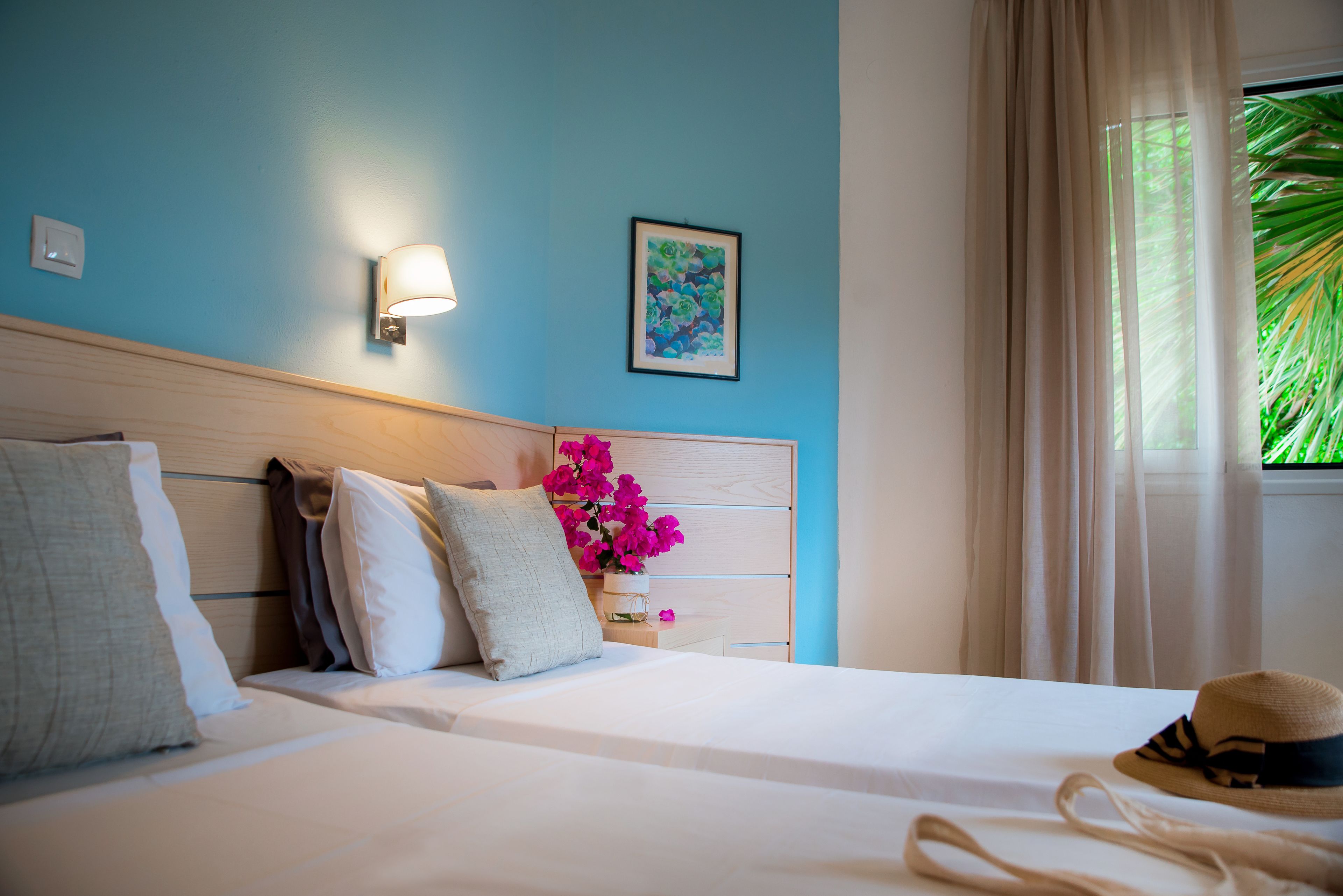 Ein Hotelzimmer mit Bett, Kissen, einem Nachttisch mit Blumenvase, einer Wandlampe und einem Fenster mit Vorhängen.