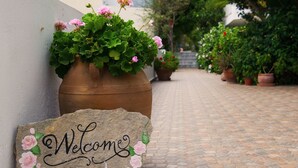 A stone welcome sign with painted roses, a potted plant with pink flowers, and a brick pathway leading to a building.