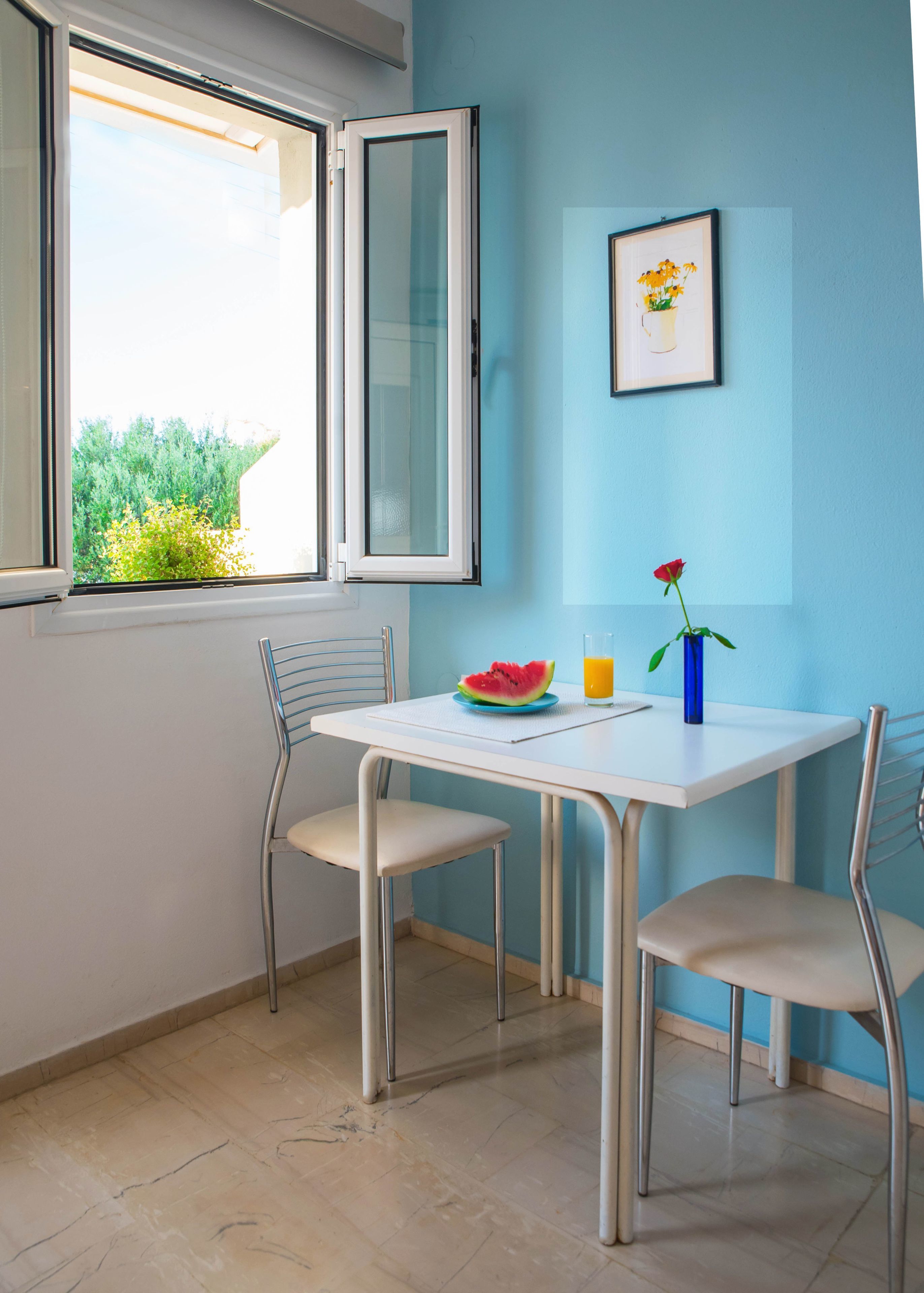 A dining area with a table, two chairs, a plate with watermelon, a glass of orange juice, and a vase with a rose.