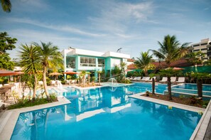 Una piscina con sillas de descanso y sombrillas, rodeada de palmeras y un edificio con grandes ventanales.