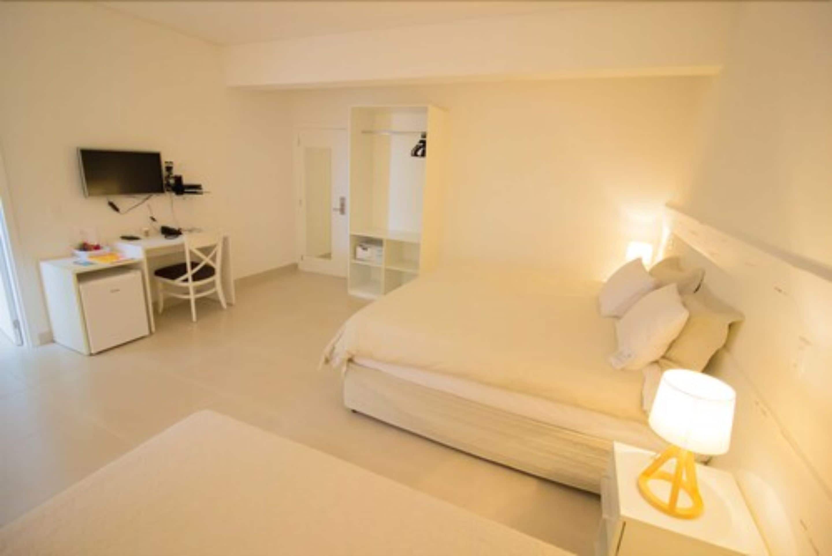 A bedroom with a bed, bedside table, lamp, desk, chair, and a TV mounted on the wall.