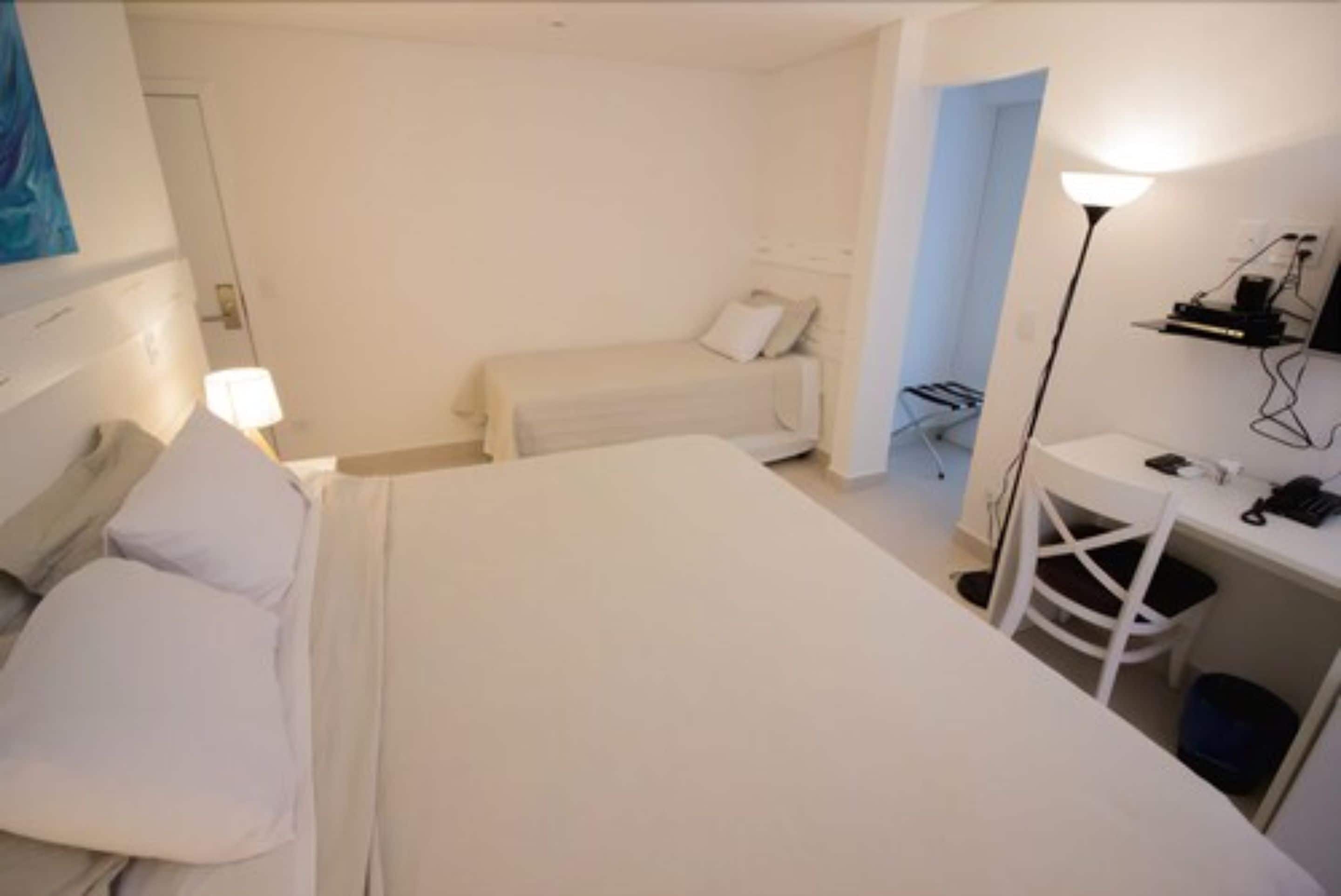 A room with a large bed, a smaller bed, a desk with a chair, a lamp, and a wall-mounted shelf.