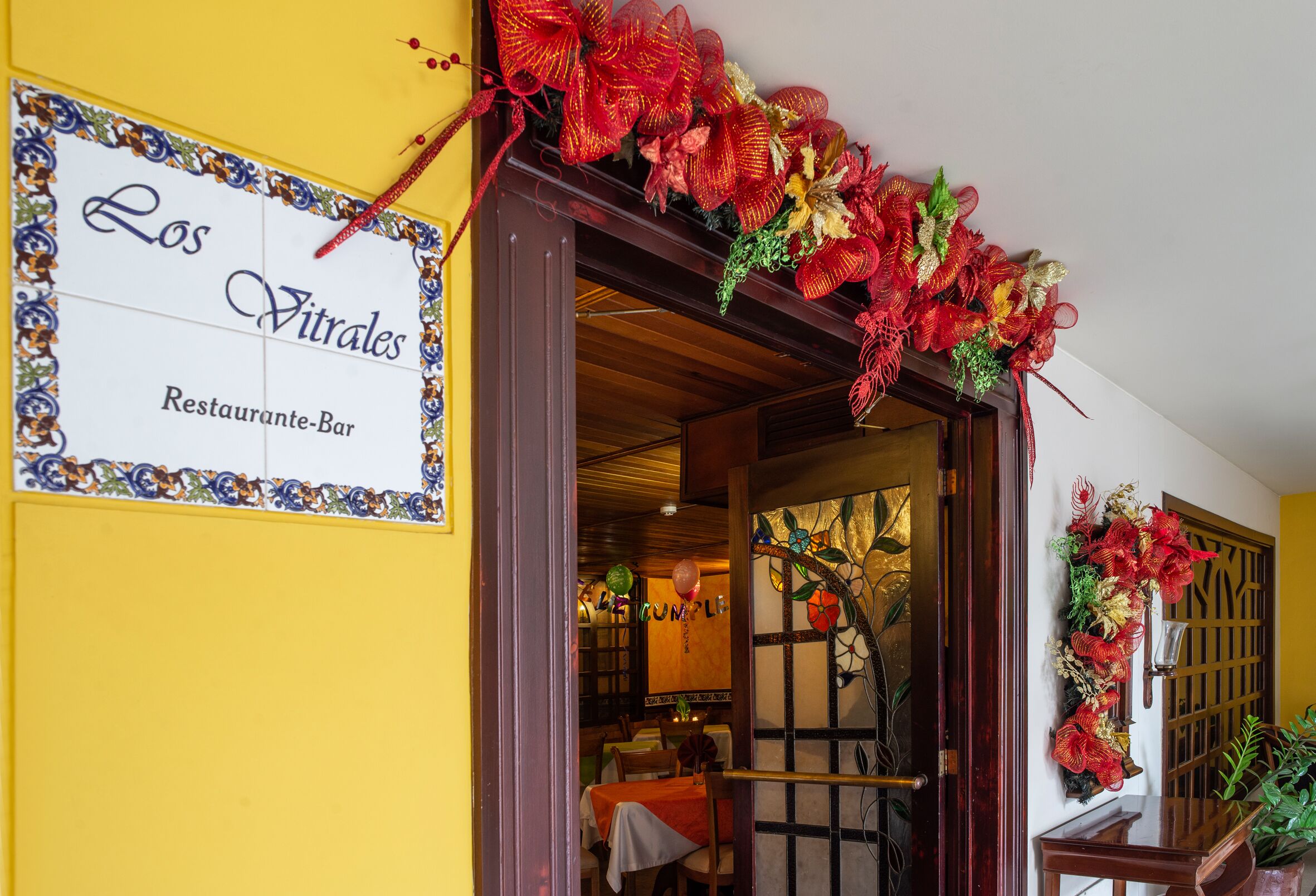 A restaurant entrance with a sign reading "Los Vitrales Restaurante-Bar" and decorative red and gold floral arrangements.