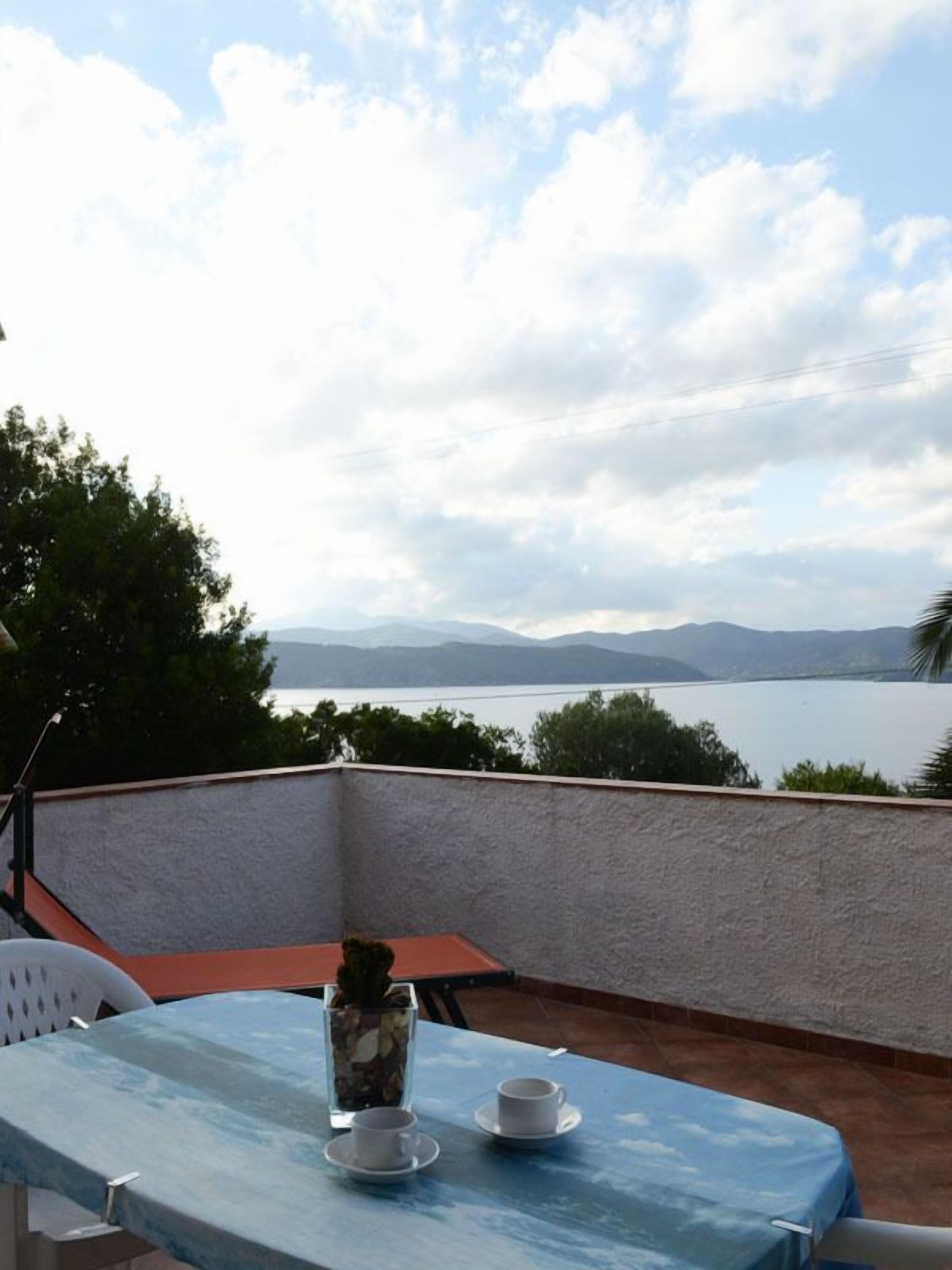 A table with two cups and a vase on a balcony overlooking a body of water and mountains.
