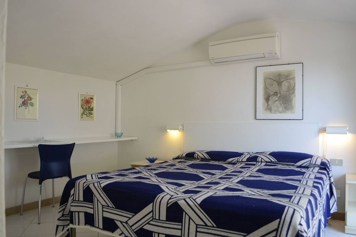 A bedroom with a bed, a desk, and a chair. There are wall-mounted lights and an air conditioner.