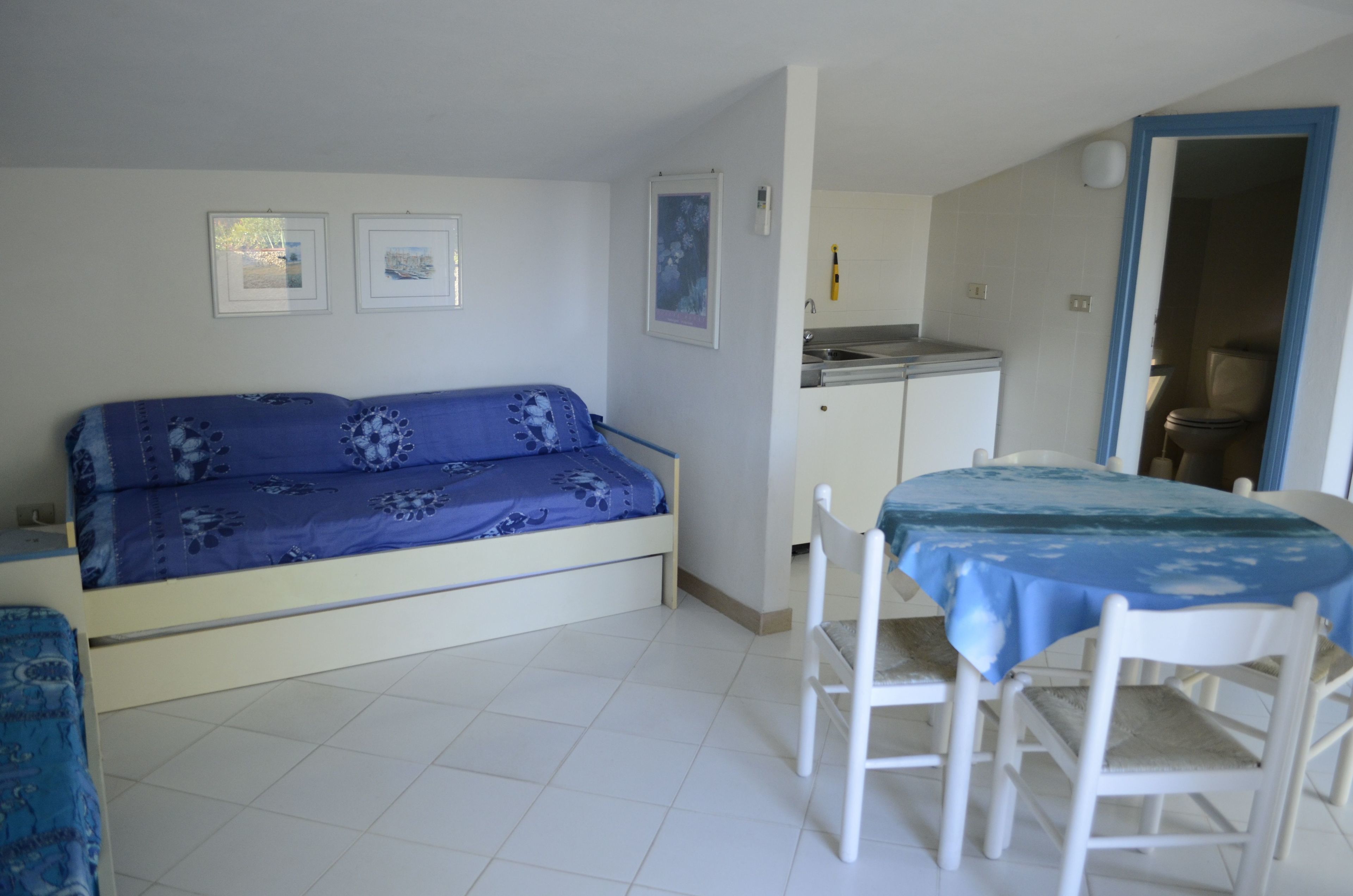 Una habitación pequeña y limpia con sofá, una mesa de comedor con sillas, un rincón americano y una puerta que da a otra habitación.