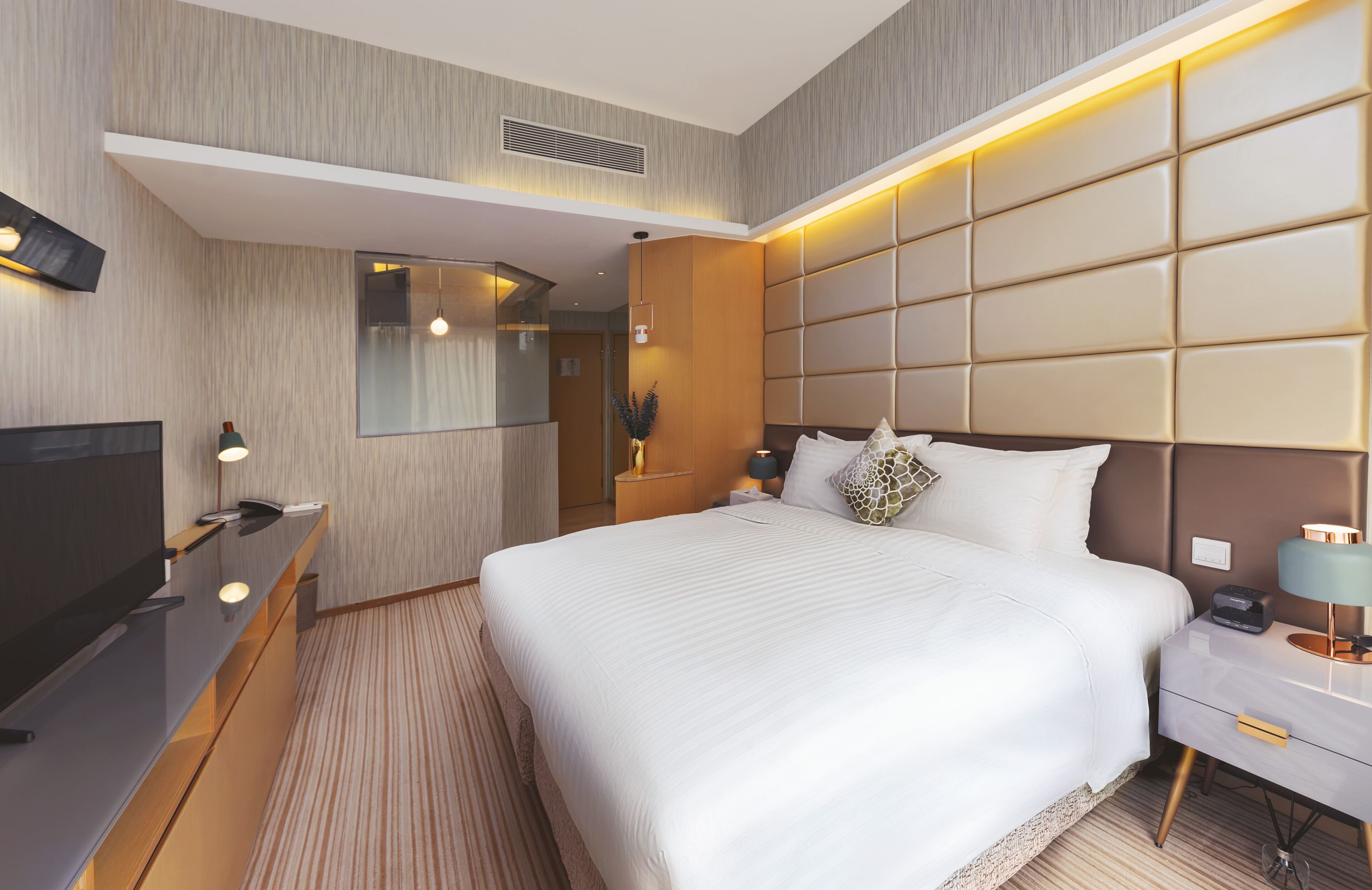 A modern hotel room with a large bed, a TV, a bedside table with a lamp, and a mirror.