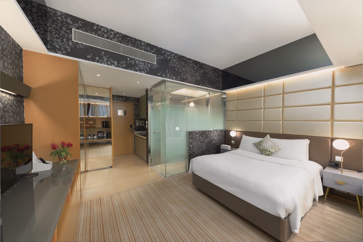 A modern hotel room with a large bed, a glass-enclosed shower, a vanity with a mirror, and a small seating area.