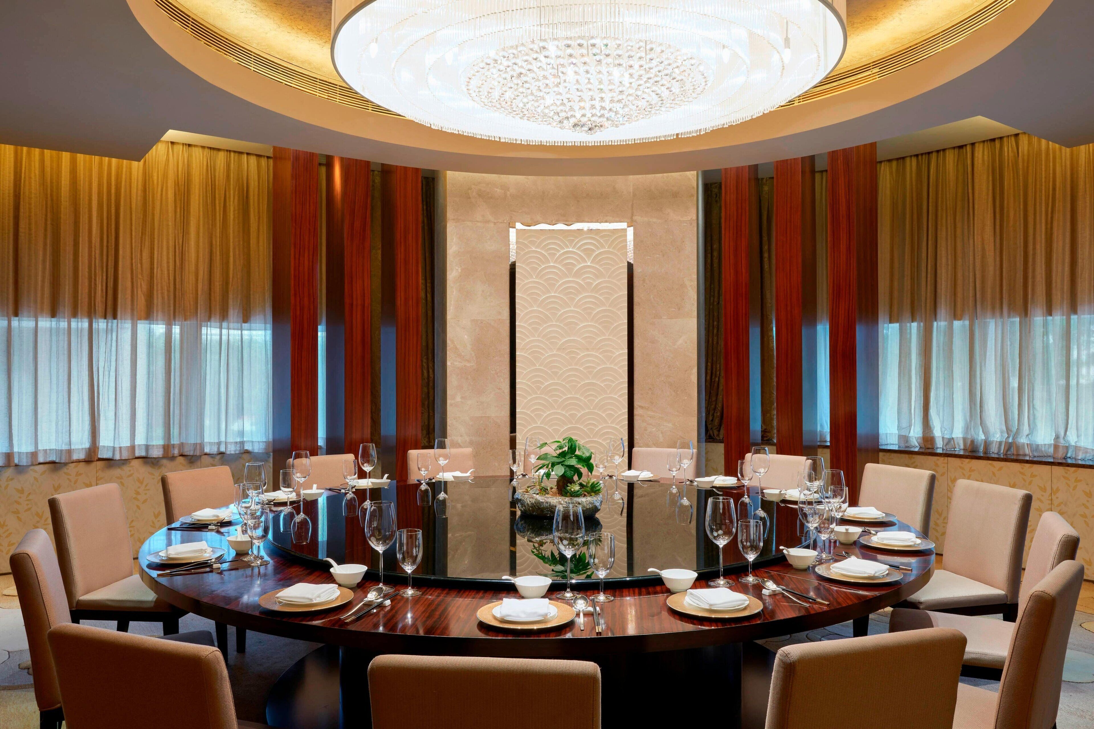 Una mesa redonda de comedor dispuesta para una comida formal, rodeada de sillas, con un candelabro sobre ella y cortinas en las ventanas.