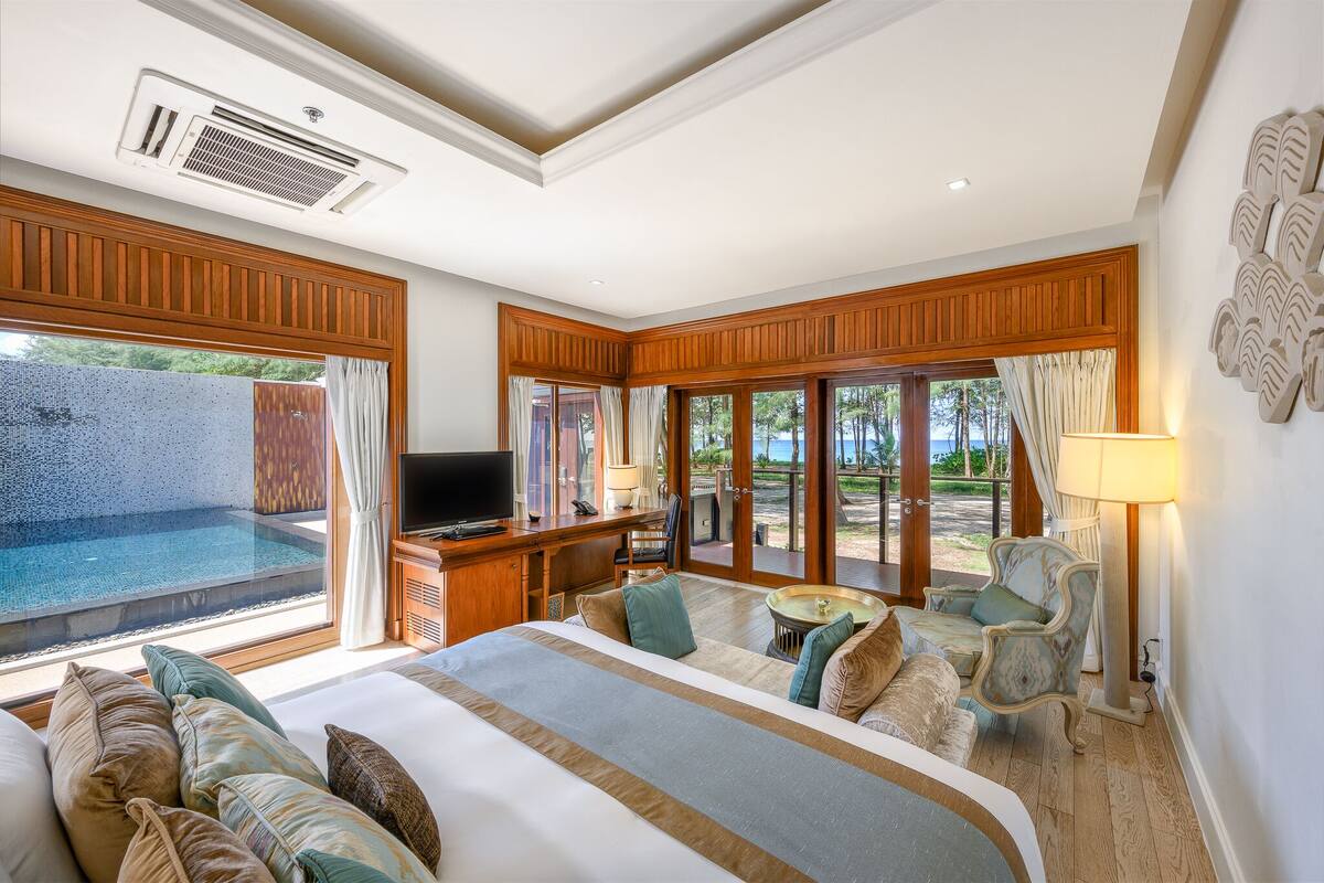 A bedroom with a large bed, a TV, a desk, a chair, a lamp, and a view of an outdoor swimming pool.